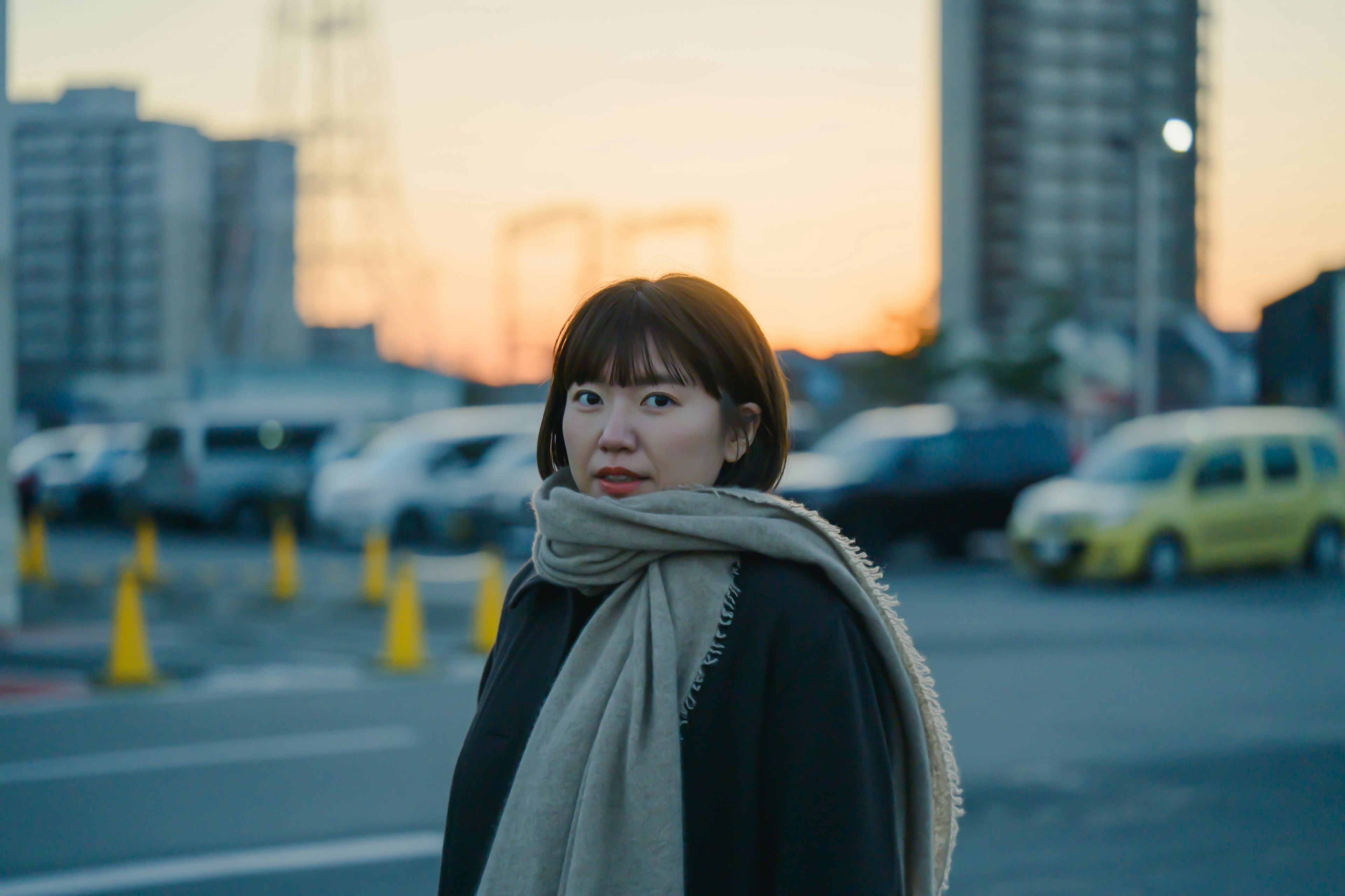 夕暮れ時に駐車場で立つ女性のポートレート 温かいスカーフを巻き込んだ黒い服を着ている 背景には都市の建物と車が見える