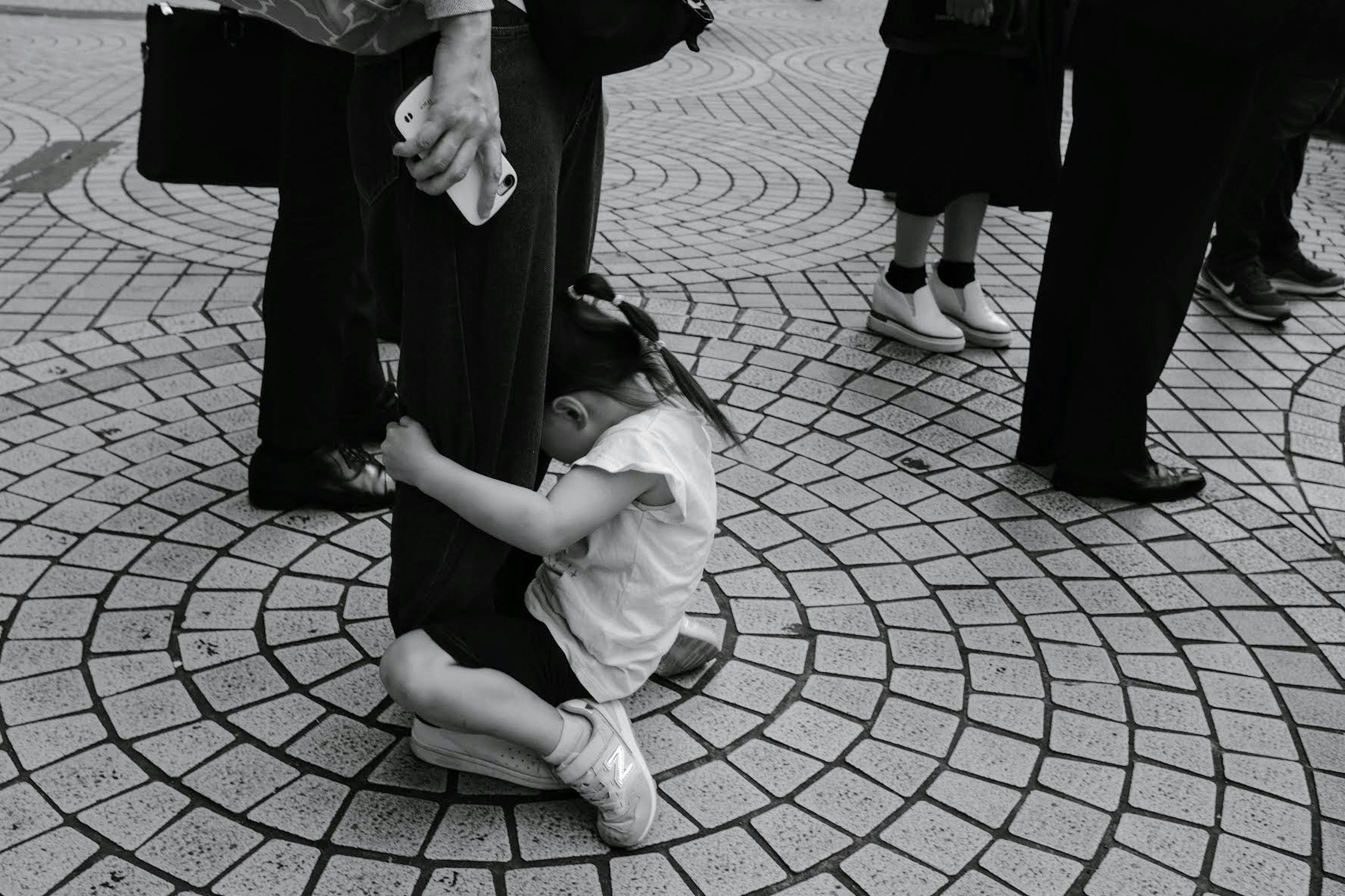 Foto hitam putih anak yang menggenggam kaki orang dewasa di lingkungan yang ramai