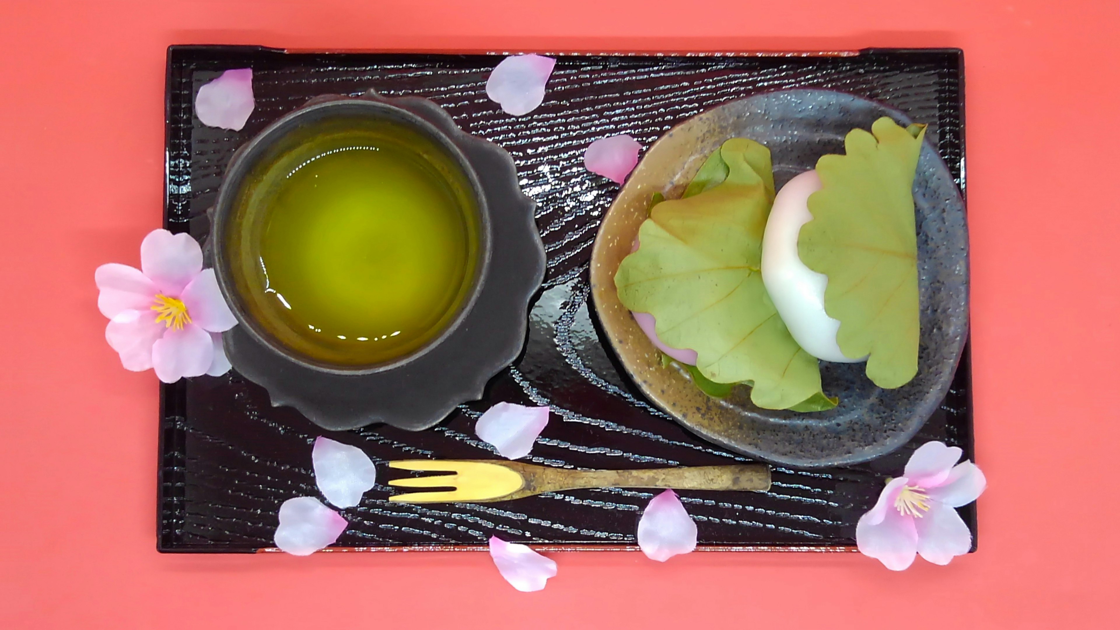 Green tea and Japanese sweets arranged on a black plate with cherry blossom petals in the background