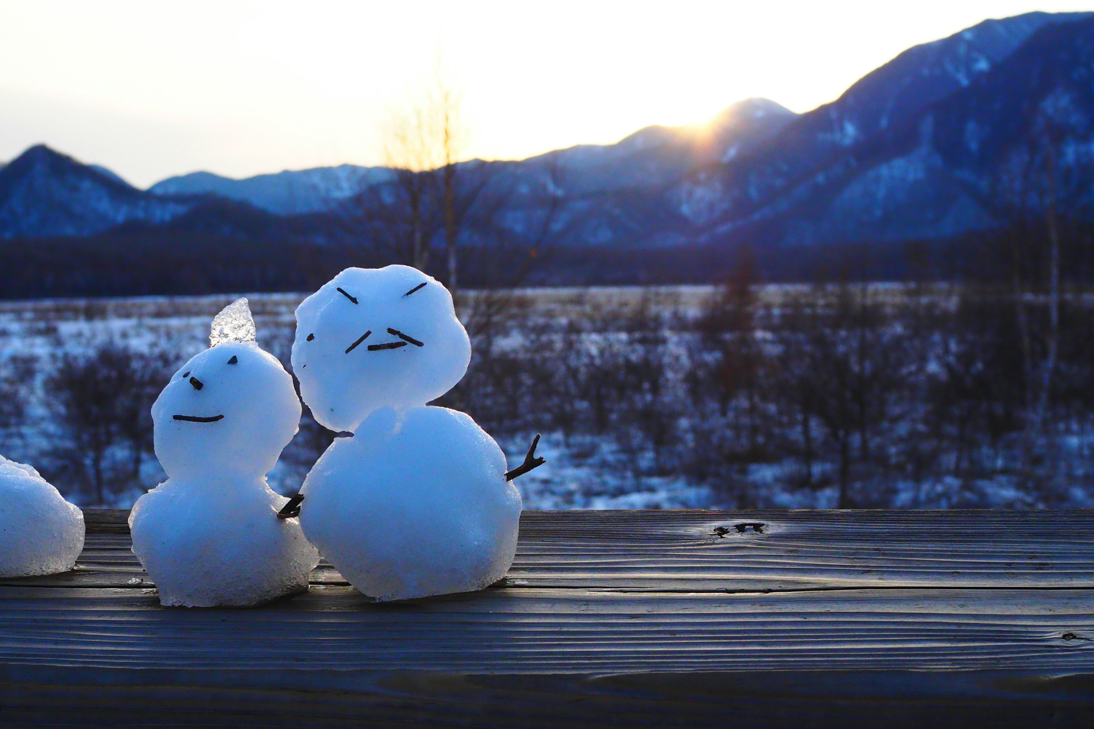 Dos muñecos de nieve sobre una superficie de madera con montañas y atardecer