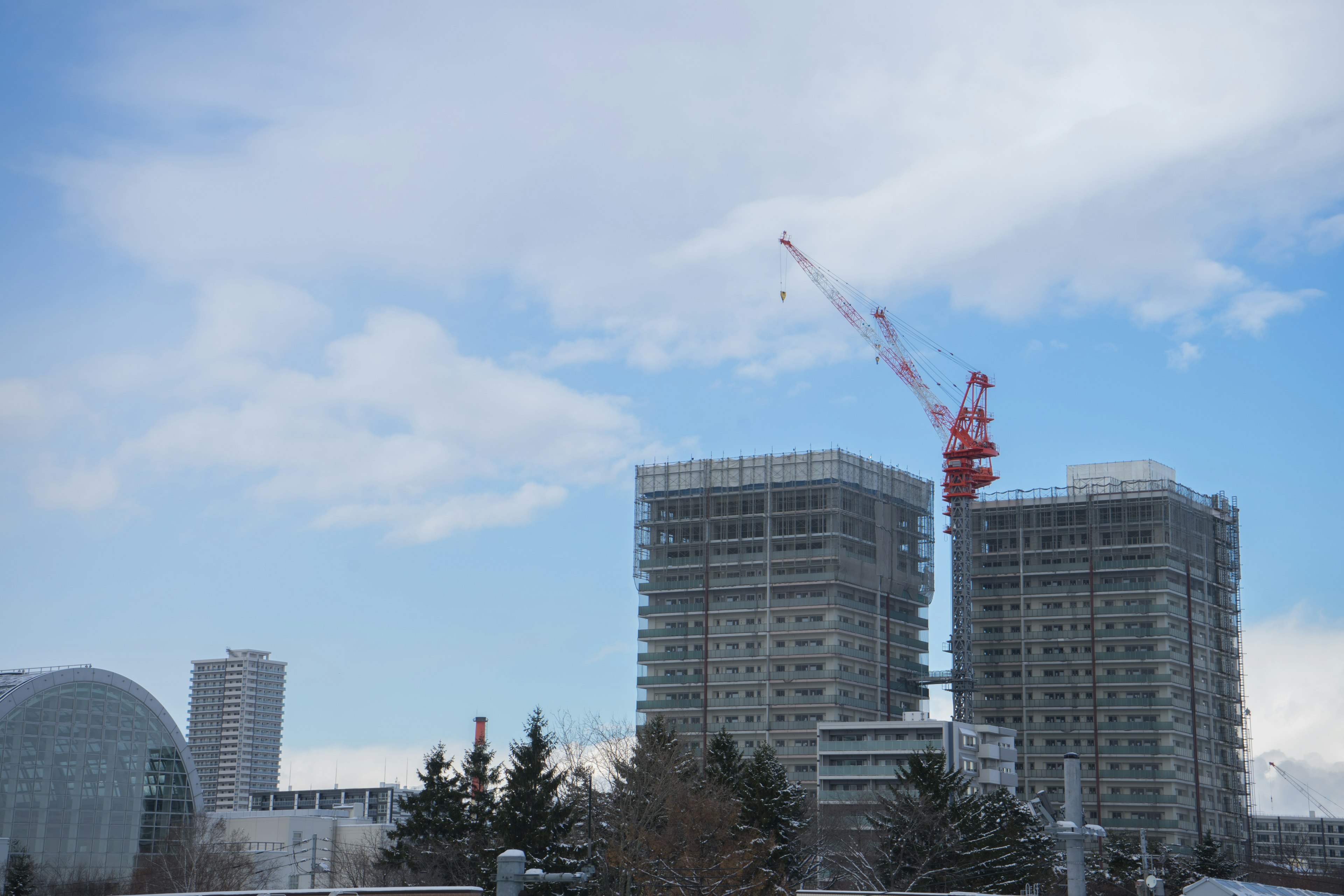 Two high-rise buildings under construction with a crane against a blue sky and white clouds