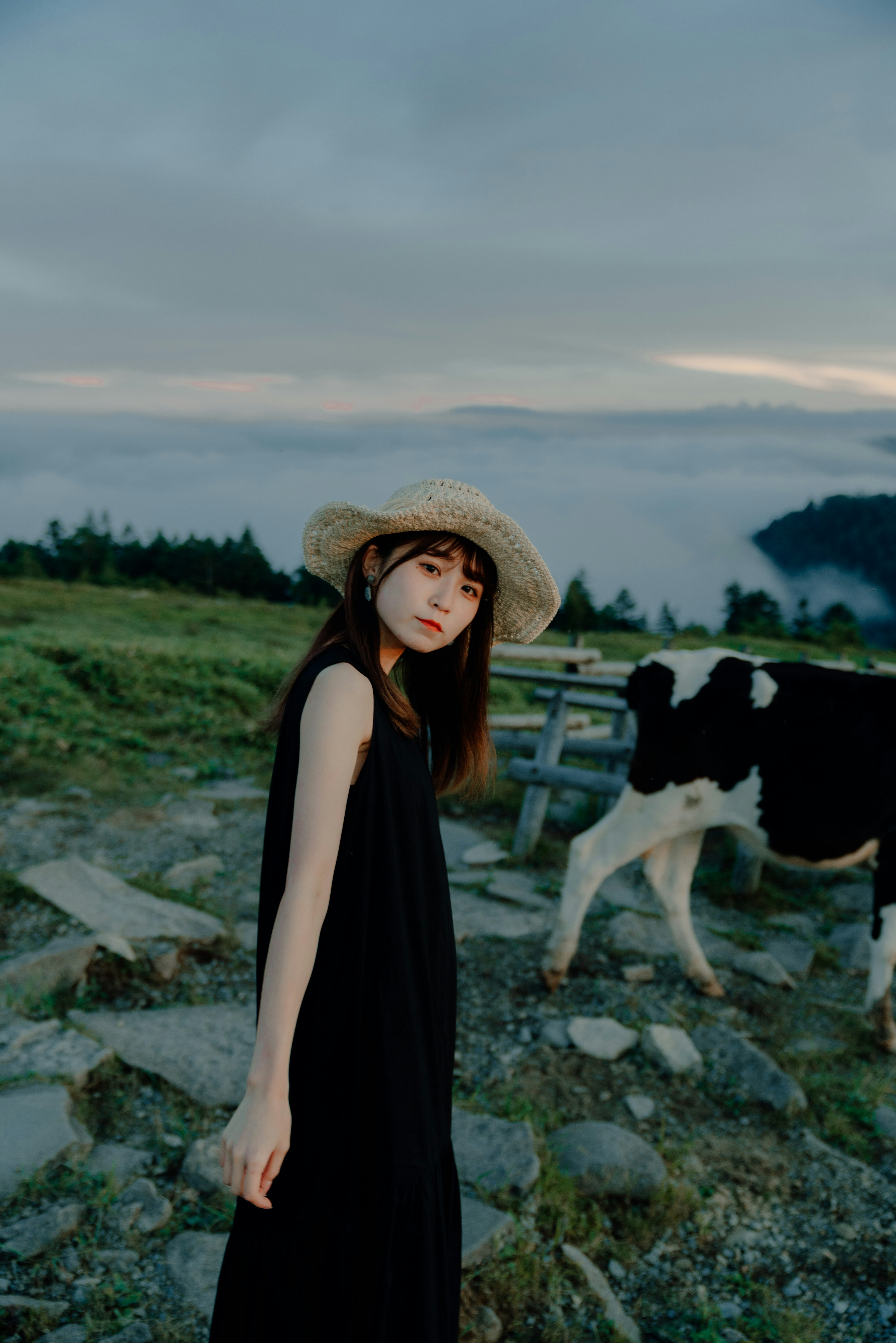 Una mujer con un vestido negro y un sombrero de paja de pie cerca de una vaca en un paisaje pintoresco