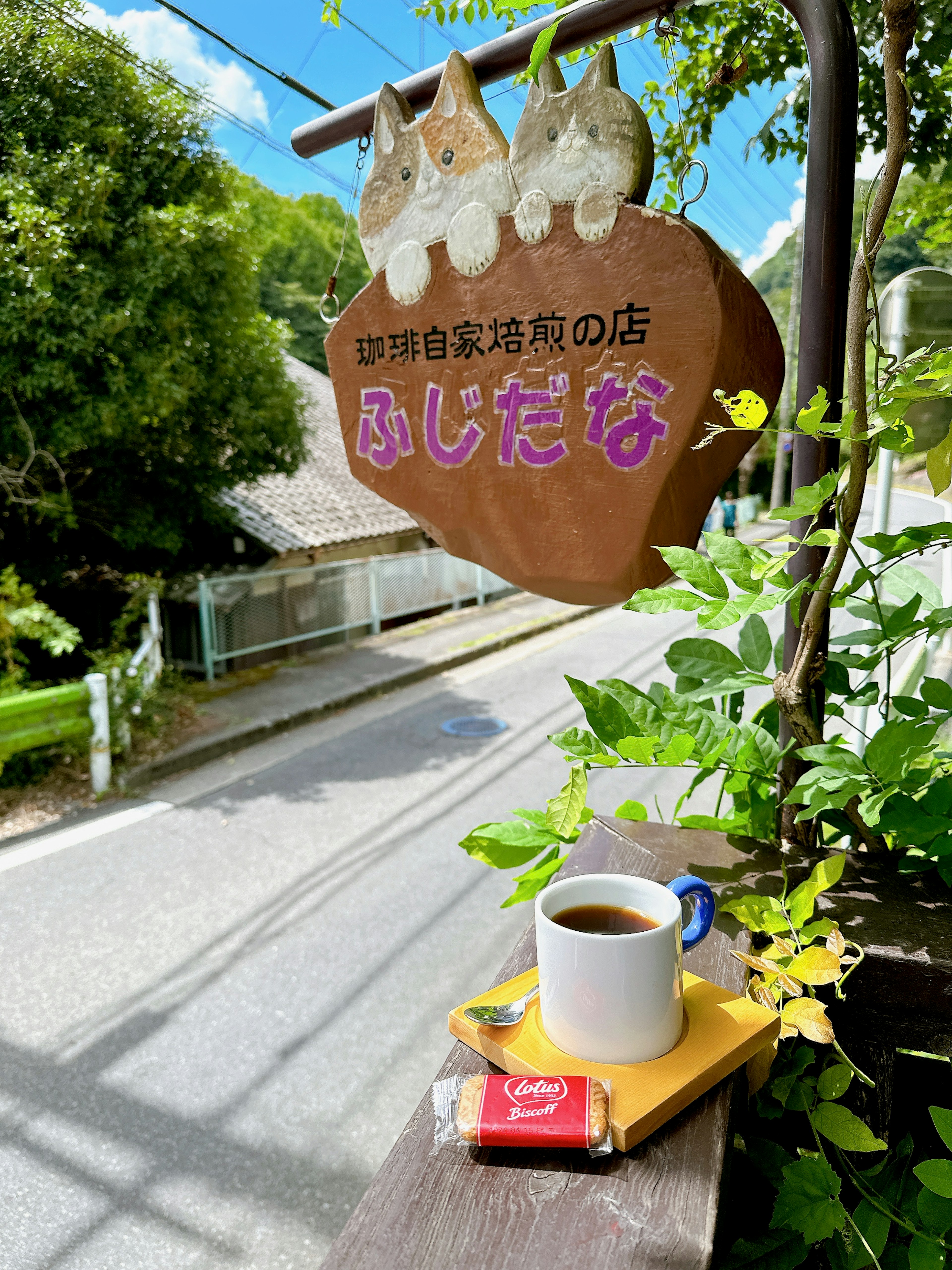 Kopi dan camilan di meja kecil kafe Papan kayu dengan nama Fujidana Tanaman hijau dan langit biru di latar belakang