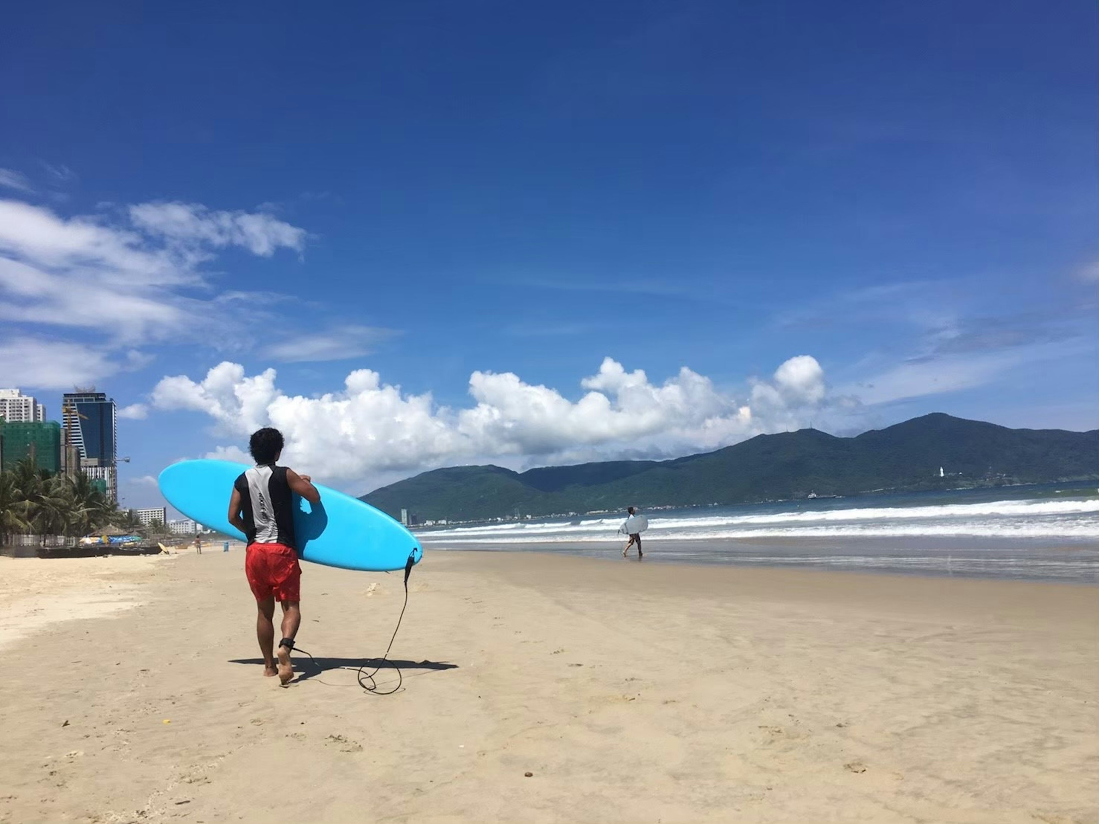 Hombre caminando por la playa sosteniendo una tabla de surf azul con cielo despejado y océano al fondo