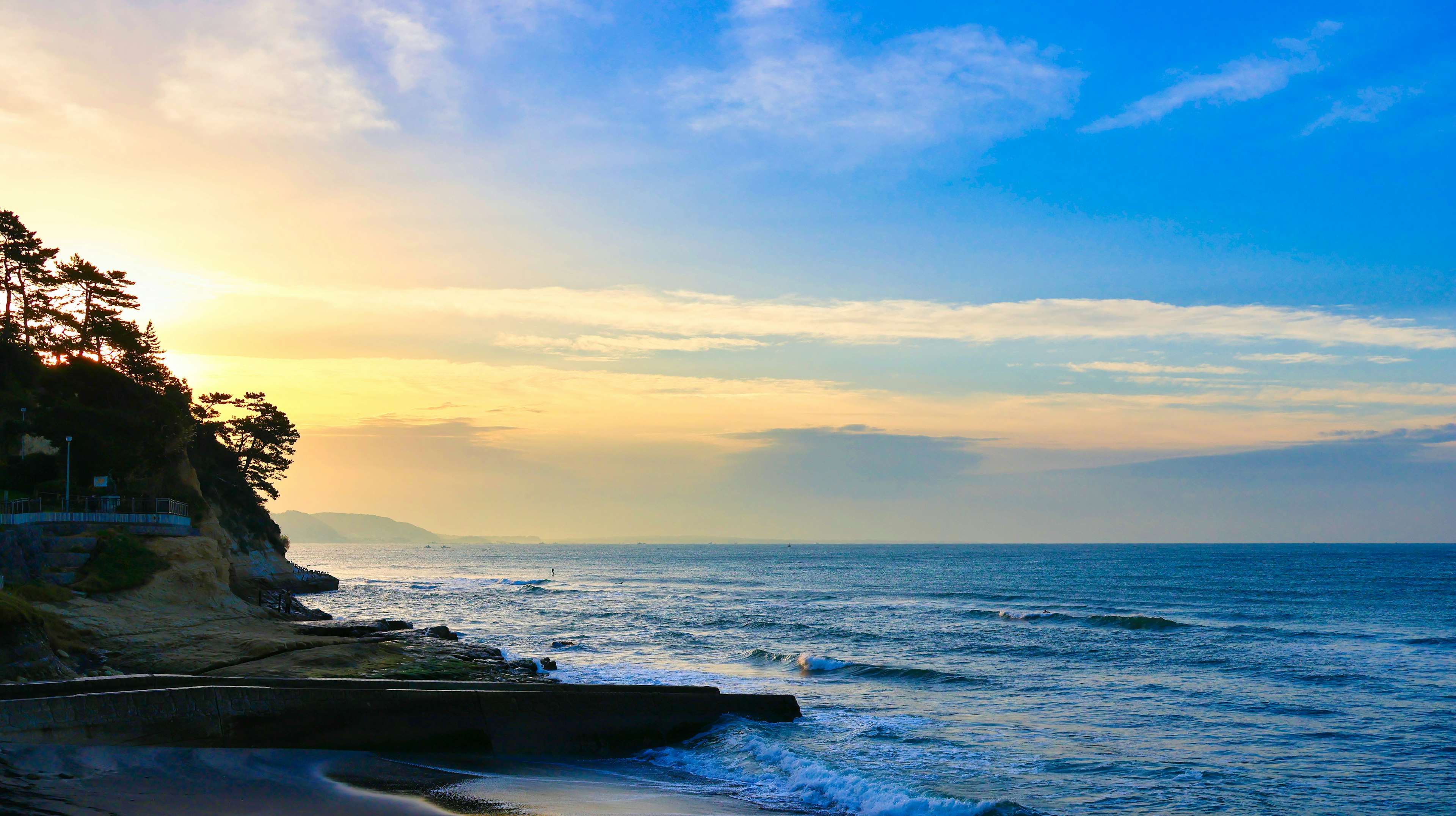 Paisaje costero al atardecer, cielo azul, olas, rocas, árboles