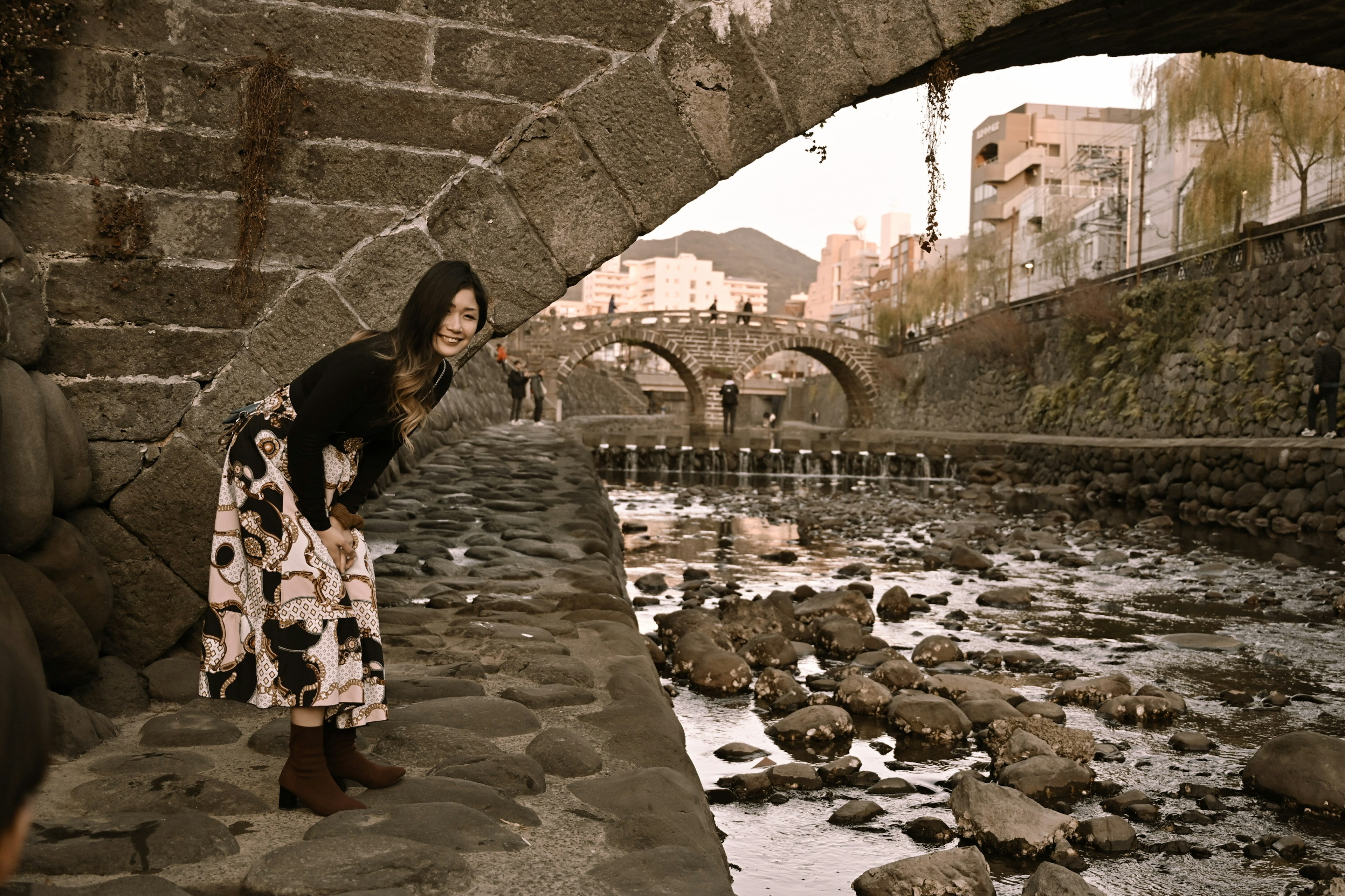 Mujer de pie junto a la orilla del río con un puente de arco de piedra al fondo