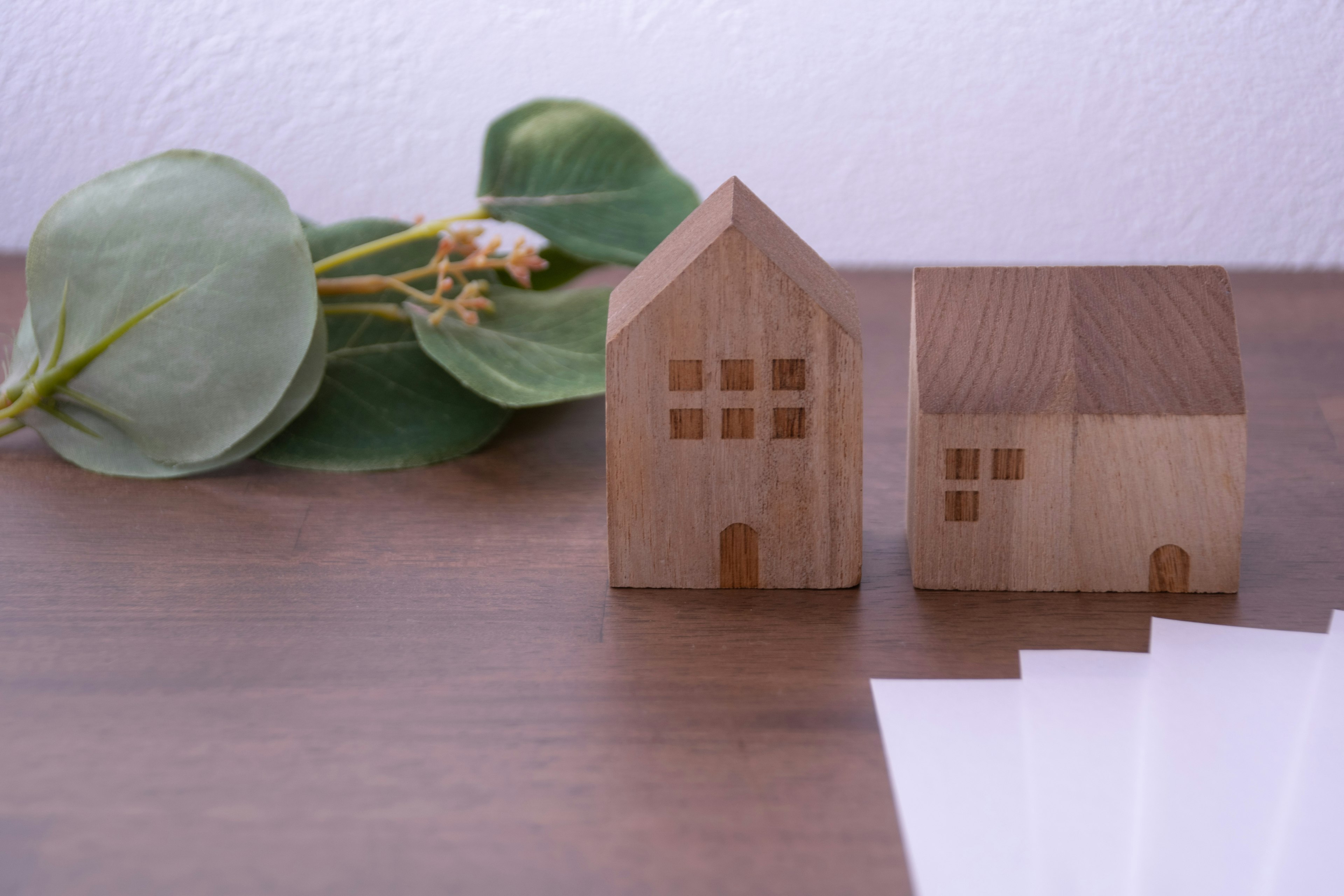 Dua model rumah kayu diletakkan di atas permukaan kayu dengan daun hijau dan kertas putih di dekatnya