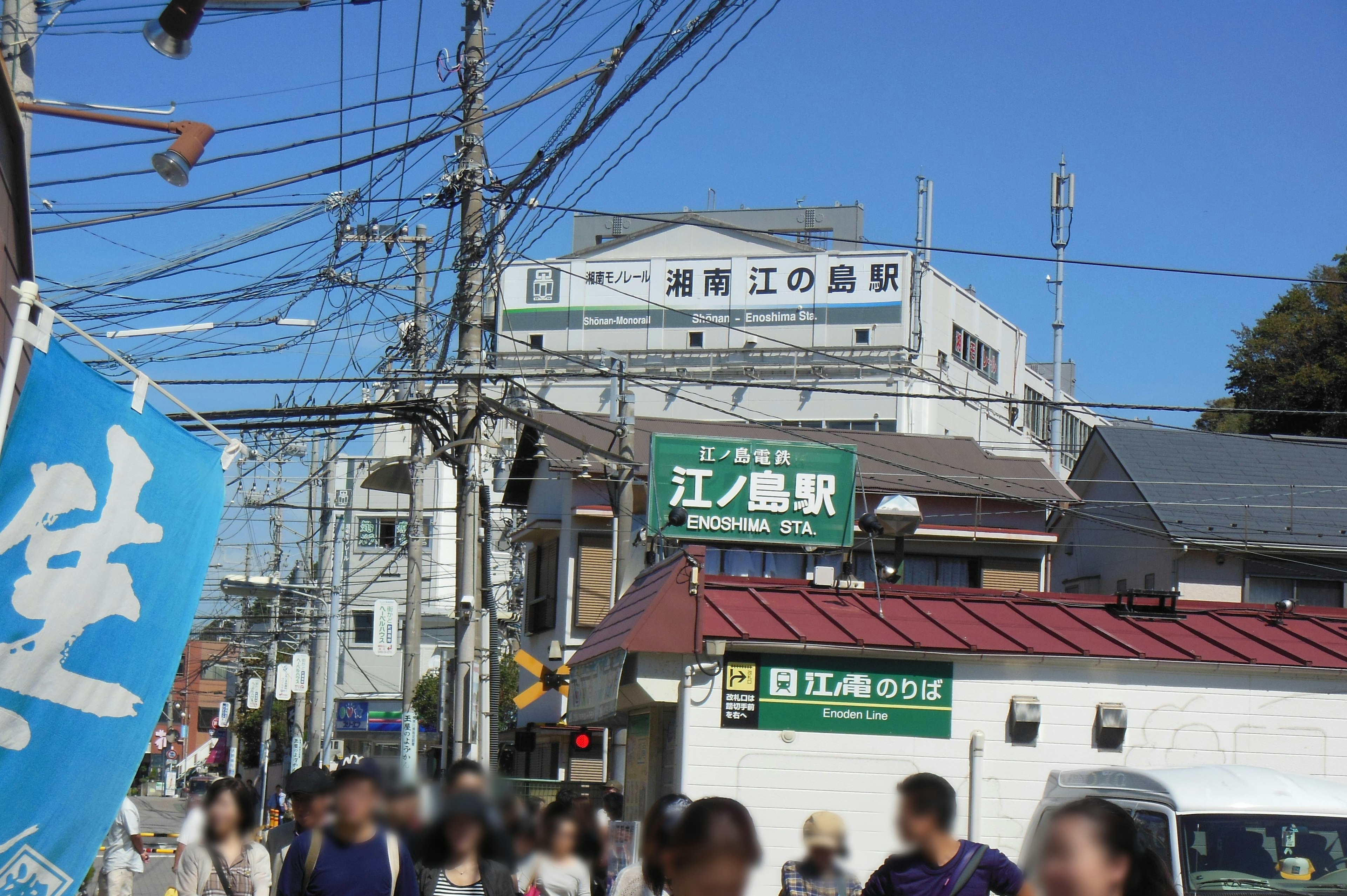 Pemandangan di area Stasiun Enoshima dengan banyak orang berjalan di bawah langit biru cerah dan saluran listrik terlihat