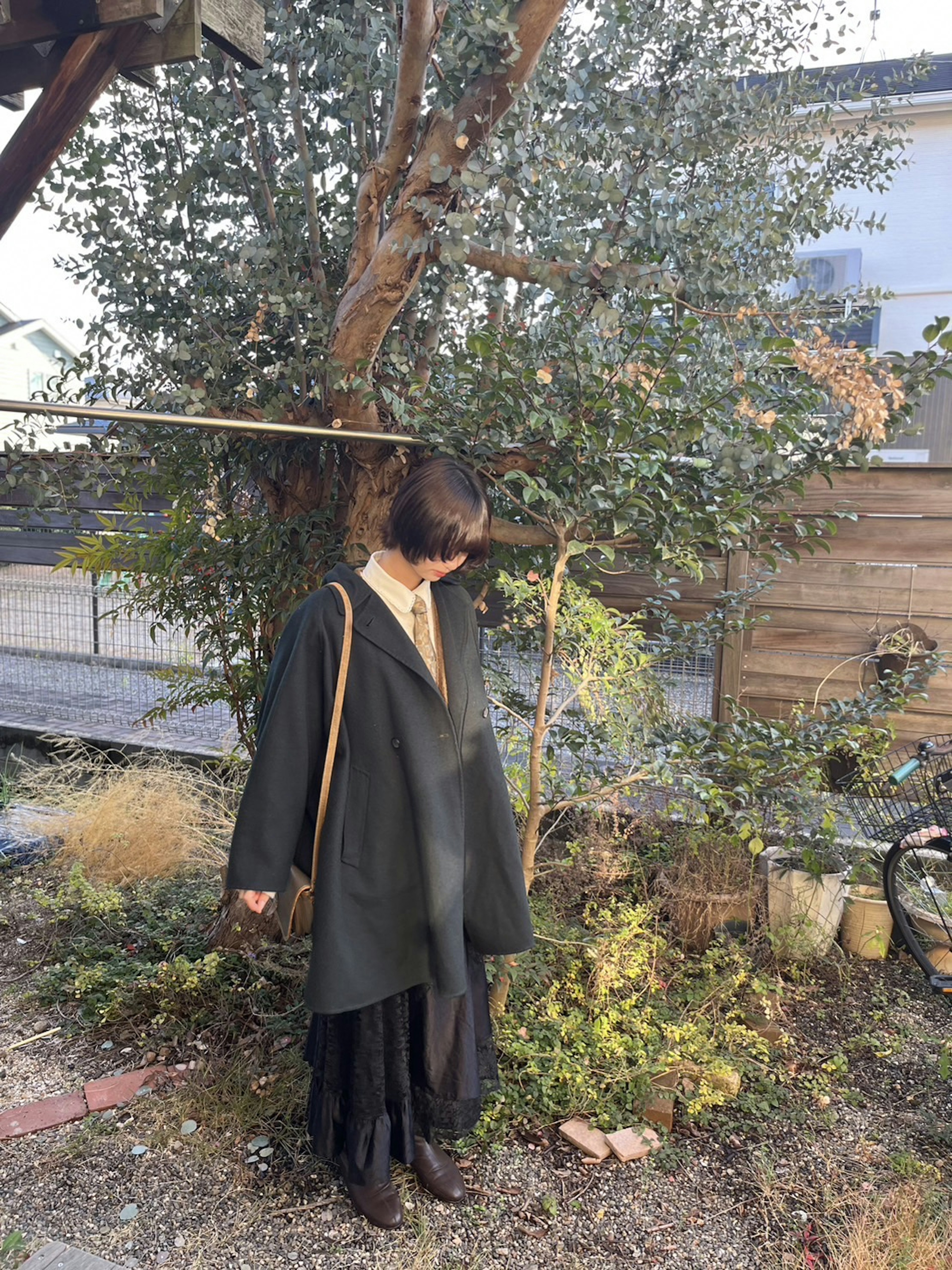 A woman wearing a green coat stands in a garden with trees and plants in the background