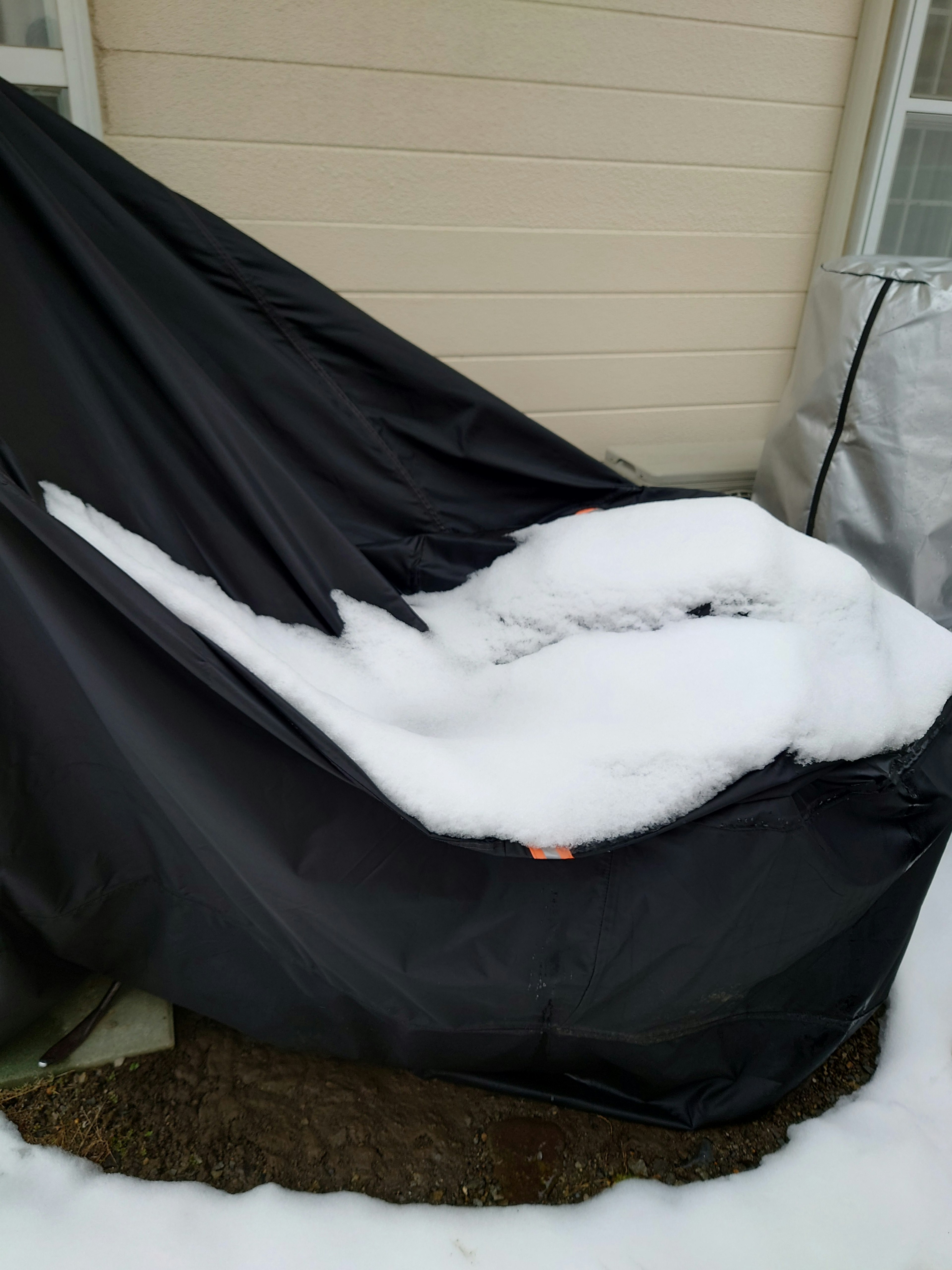 Snow covering a black outdoor cover with a beige wall in the background