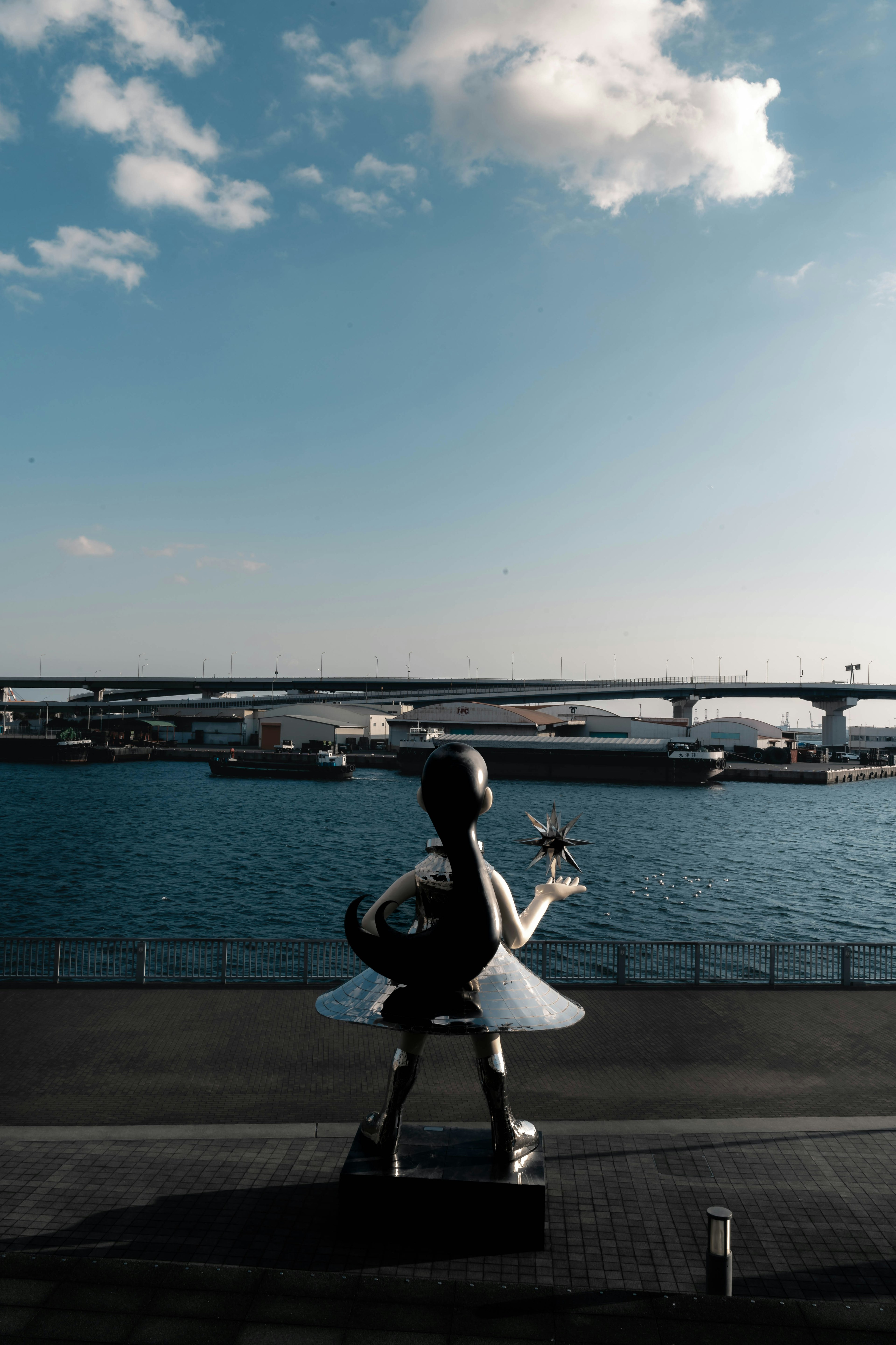 Una estatua de bailarina de espaldas al mar con un puente al fondo