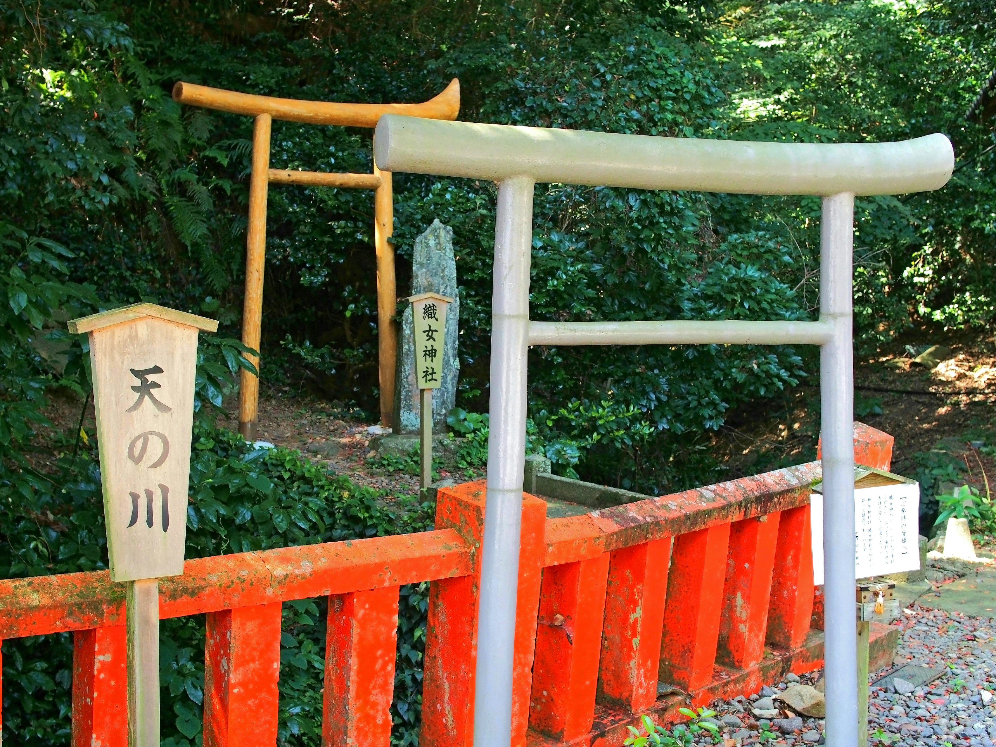 A serene scene featuring two torii gates surrounded by greenery One gate is orange and the other is grey stone with a river nearby