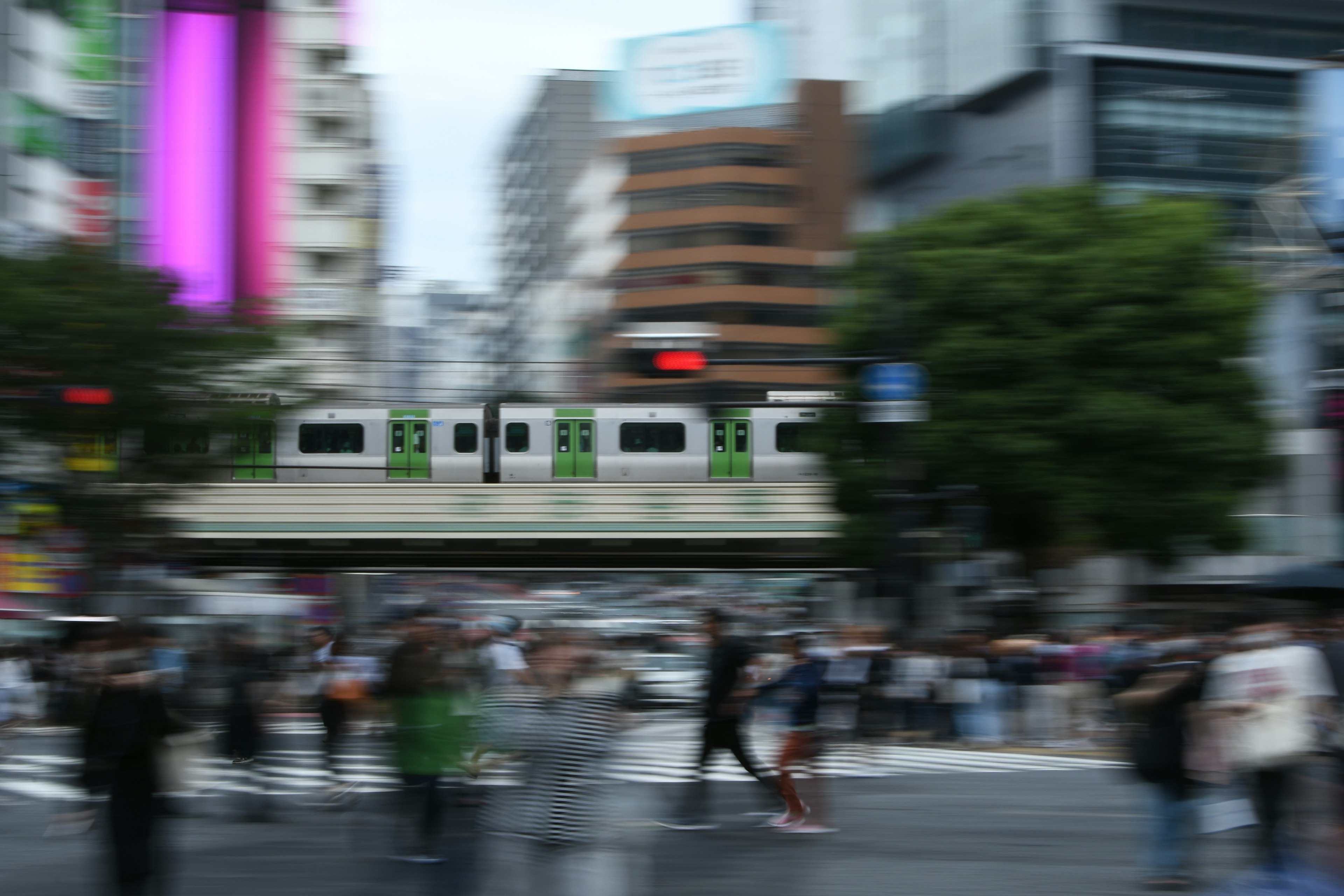 Blurred urban intersection with people crossing and a green train above