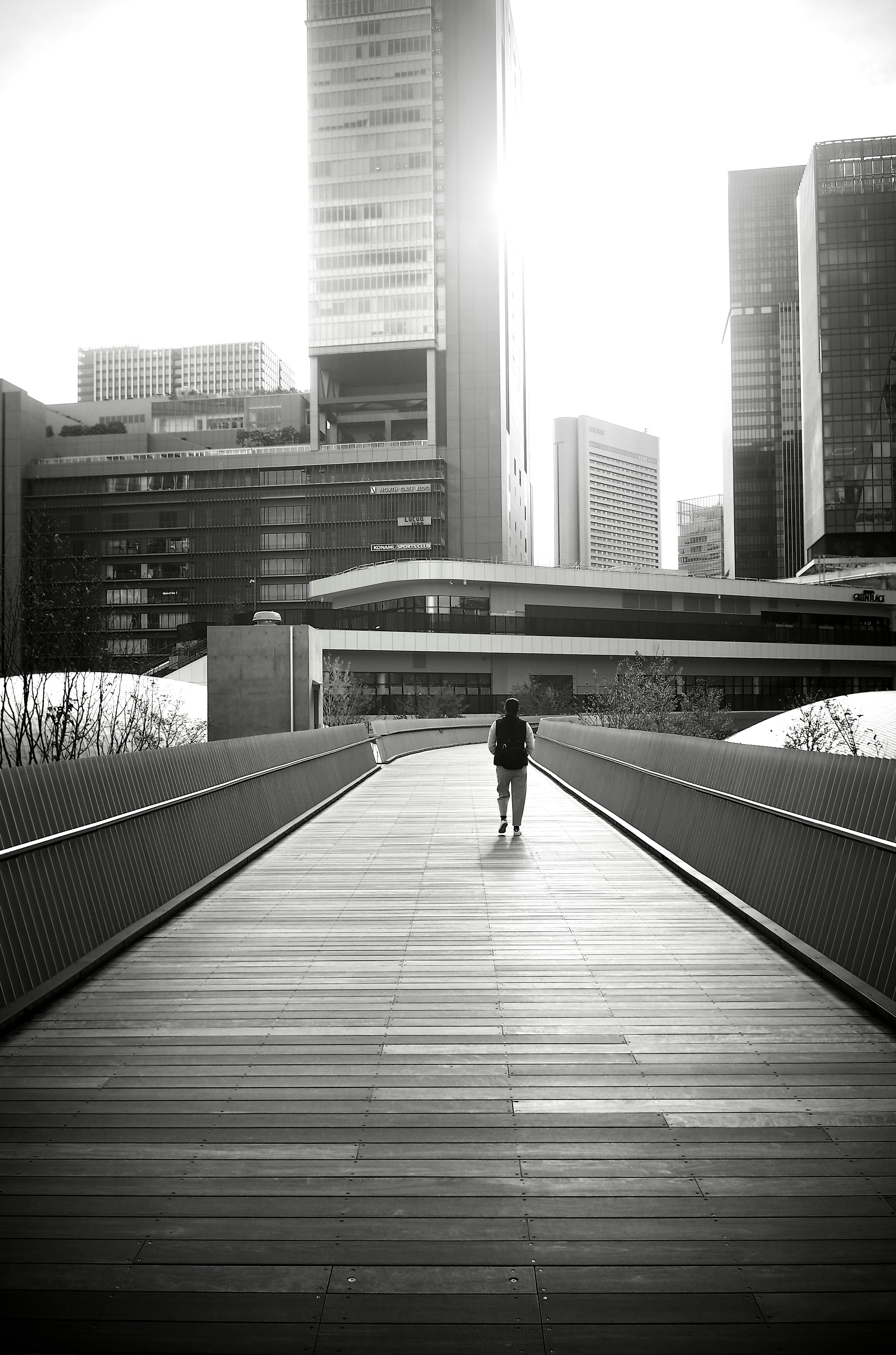 Black and white photo of a person walking on a wooden pathway surrounded by skyscrapers