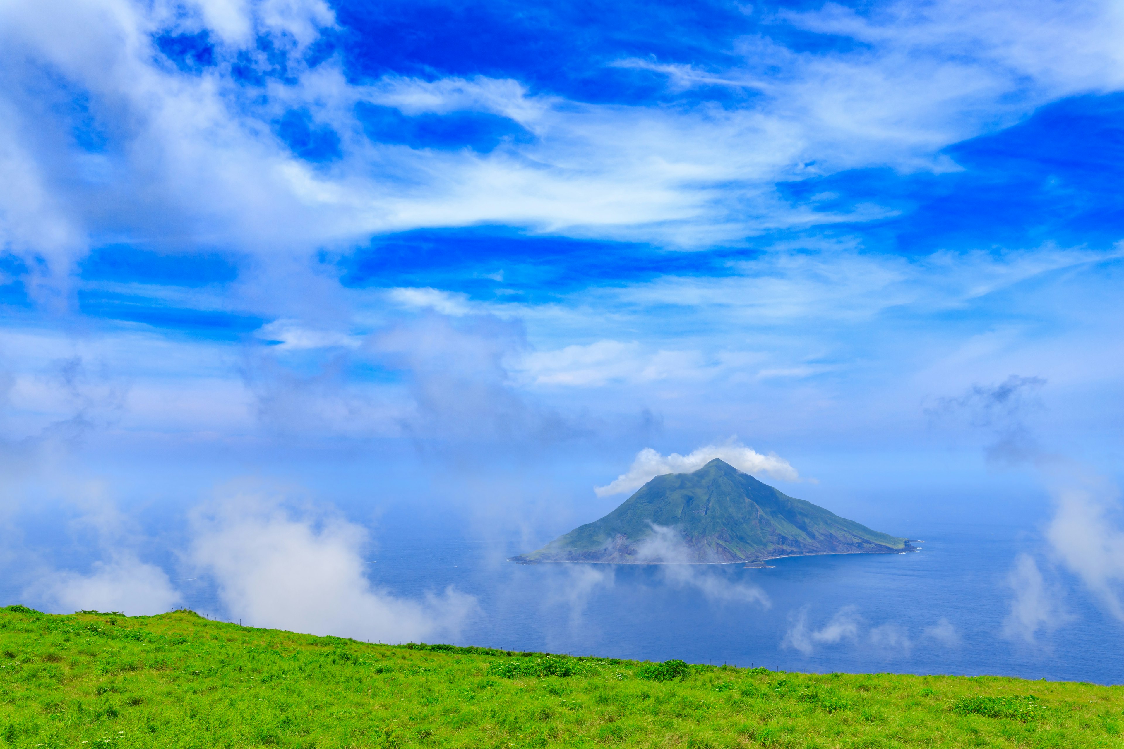 Ein grüner Hügel unter einem blauen Himmel mit Wolken und einer kleinen Insel im Ozean