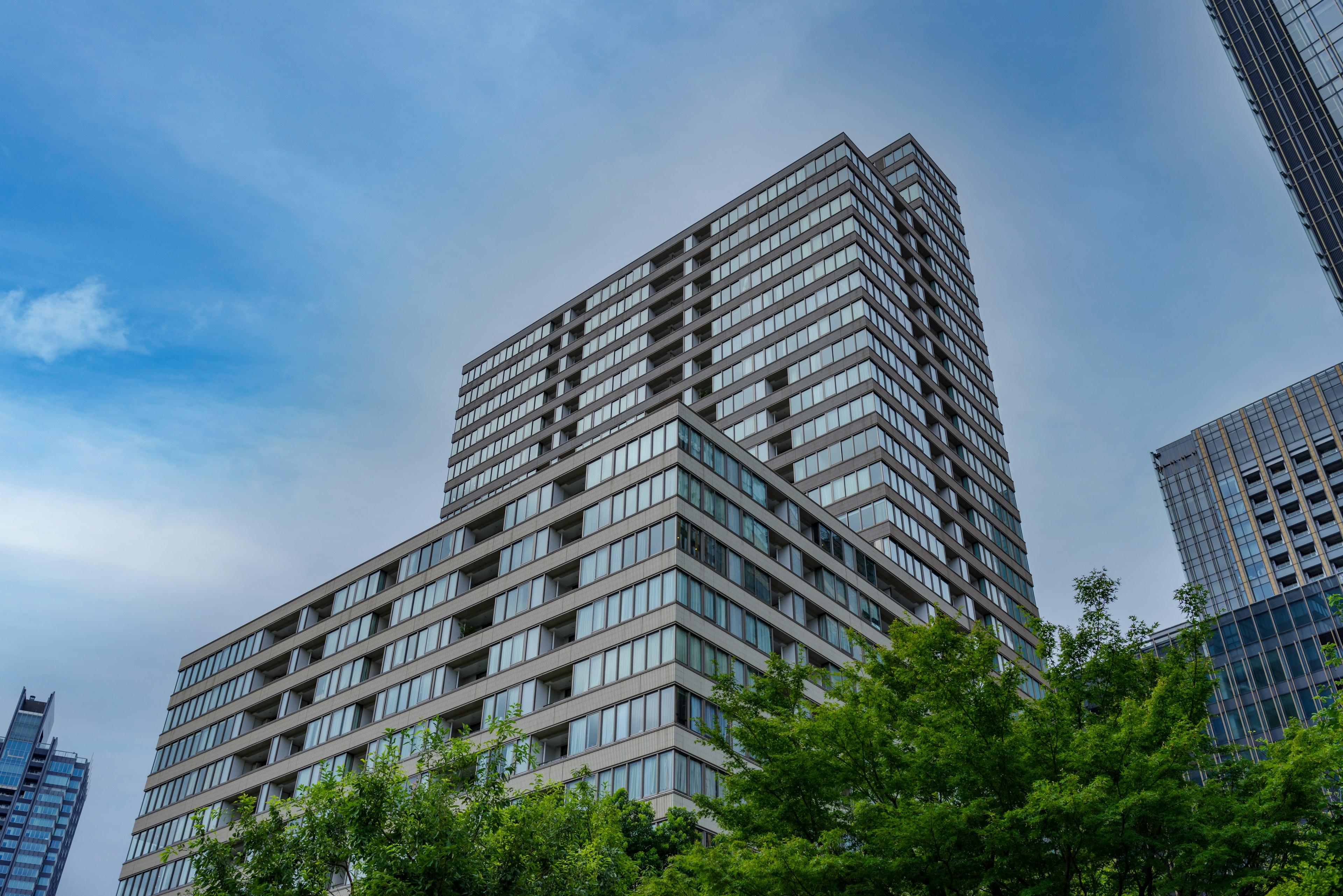 Close-up of a high-rise building with glass facade under a blue sky and clouds surrounded by green trees