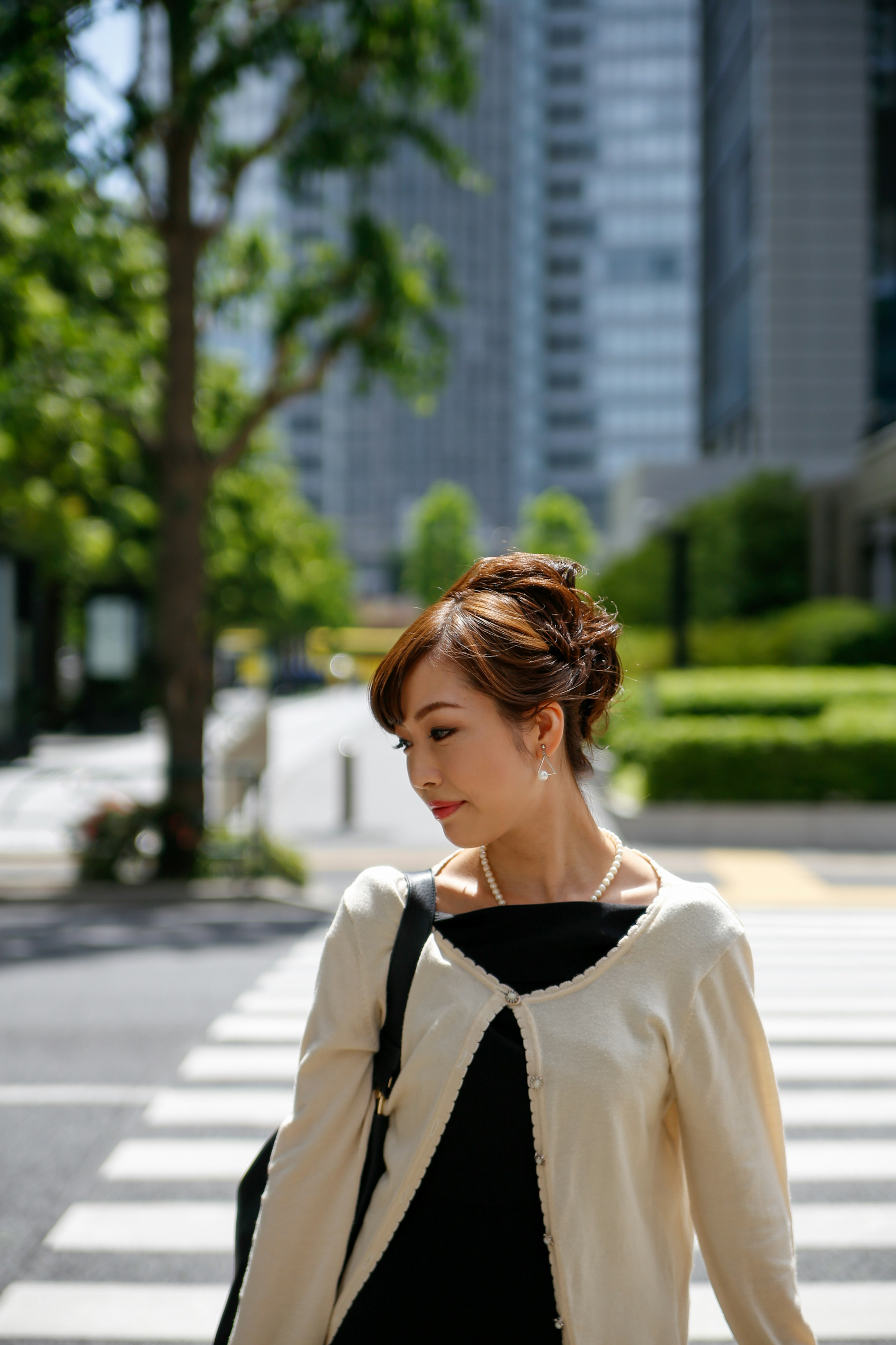 Woman turning her head on an urban sidewalk wearing an off-white cardigan and black dress surrounded by tall buildings and green trees