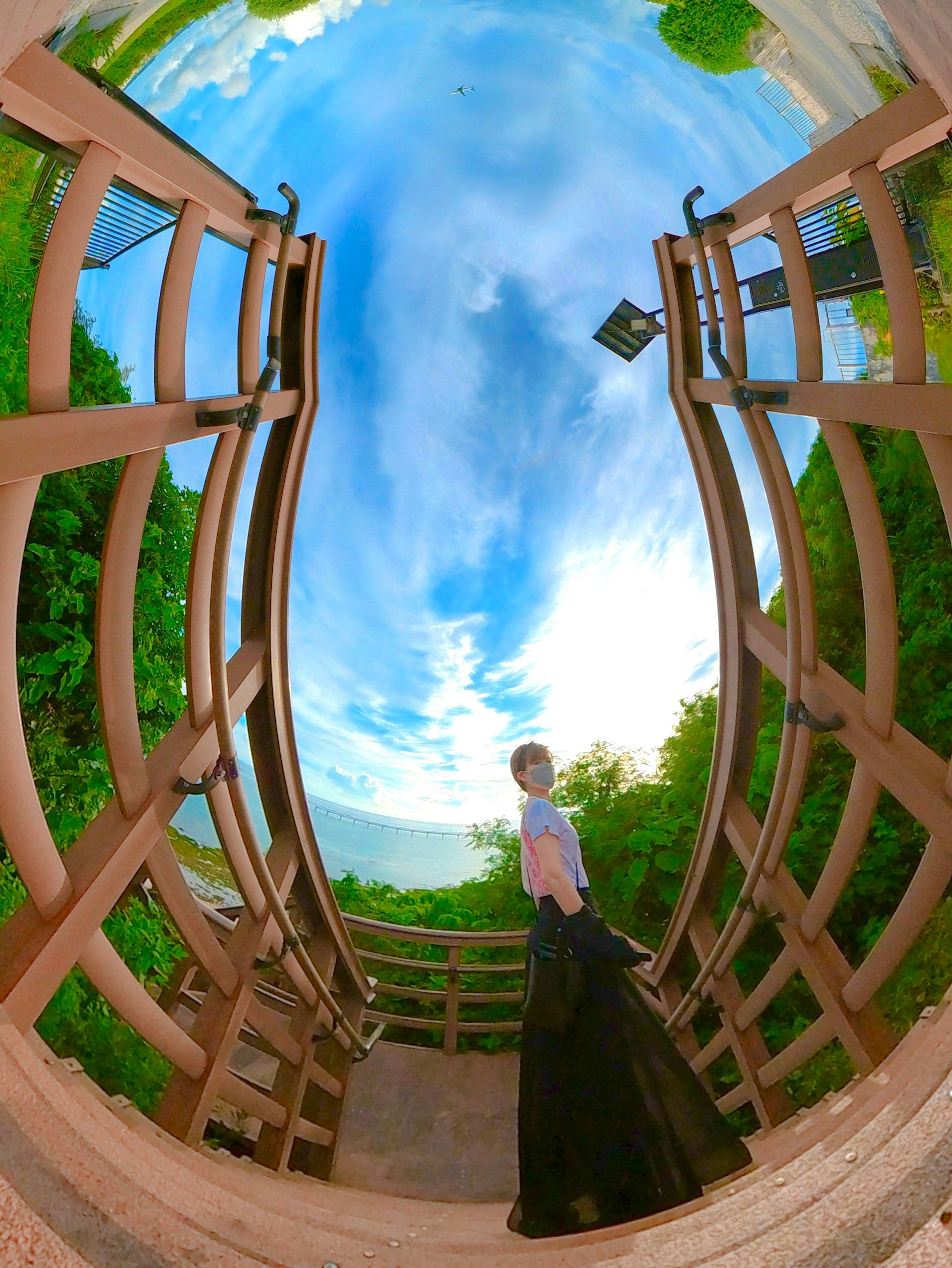 A woman standing on a wooden bridge with a blue sky and greenery in the background