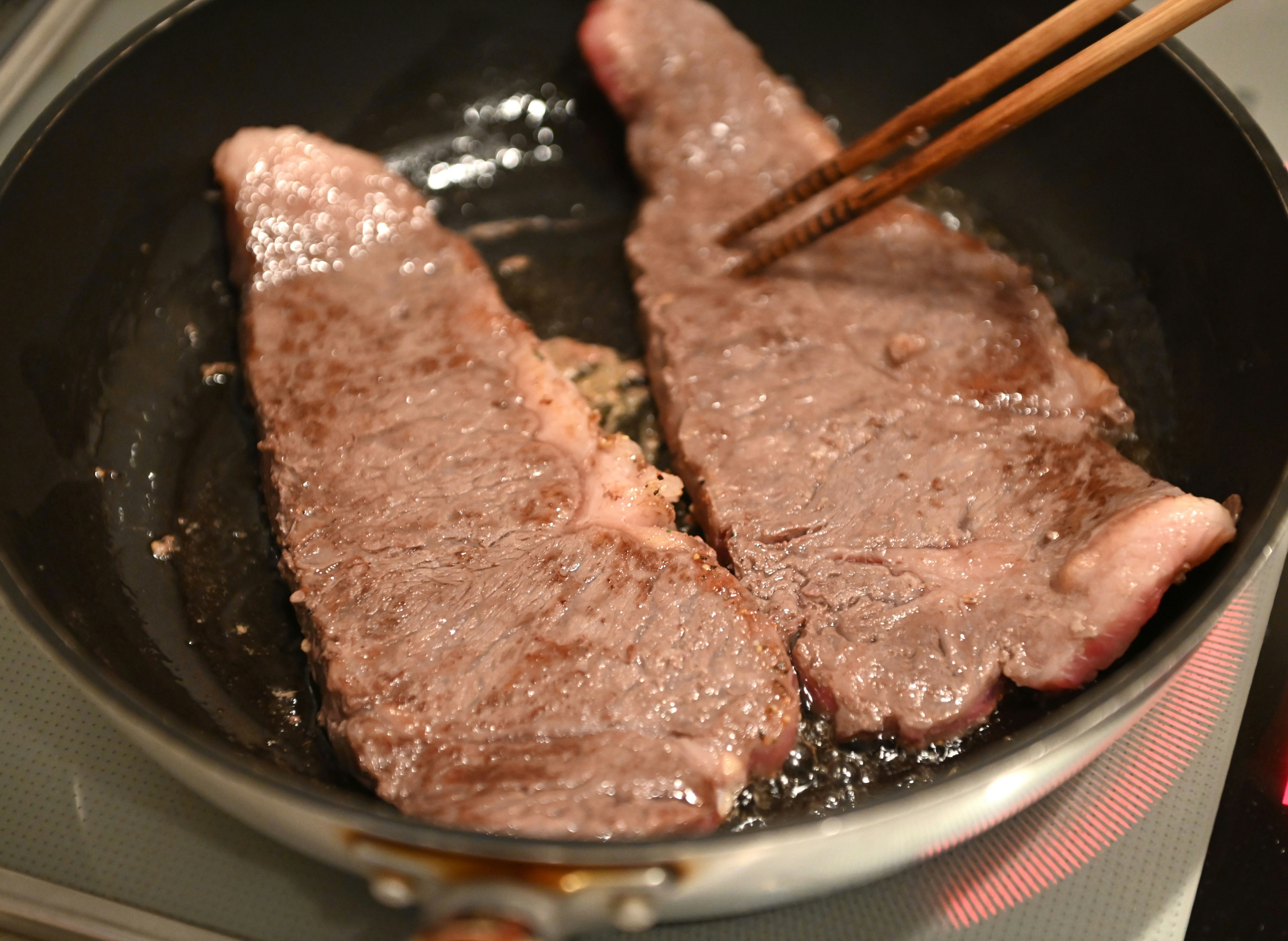 Two beef steaks cooking in a frying pan with chopsticks