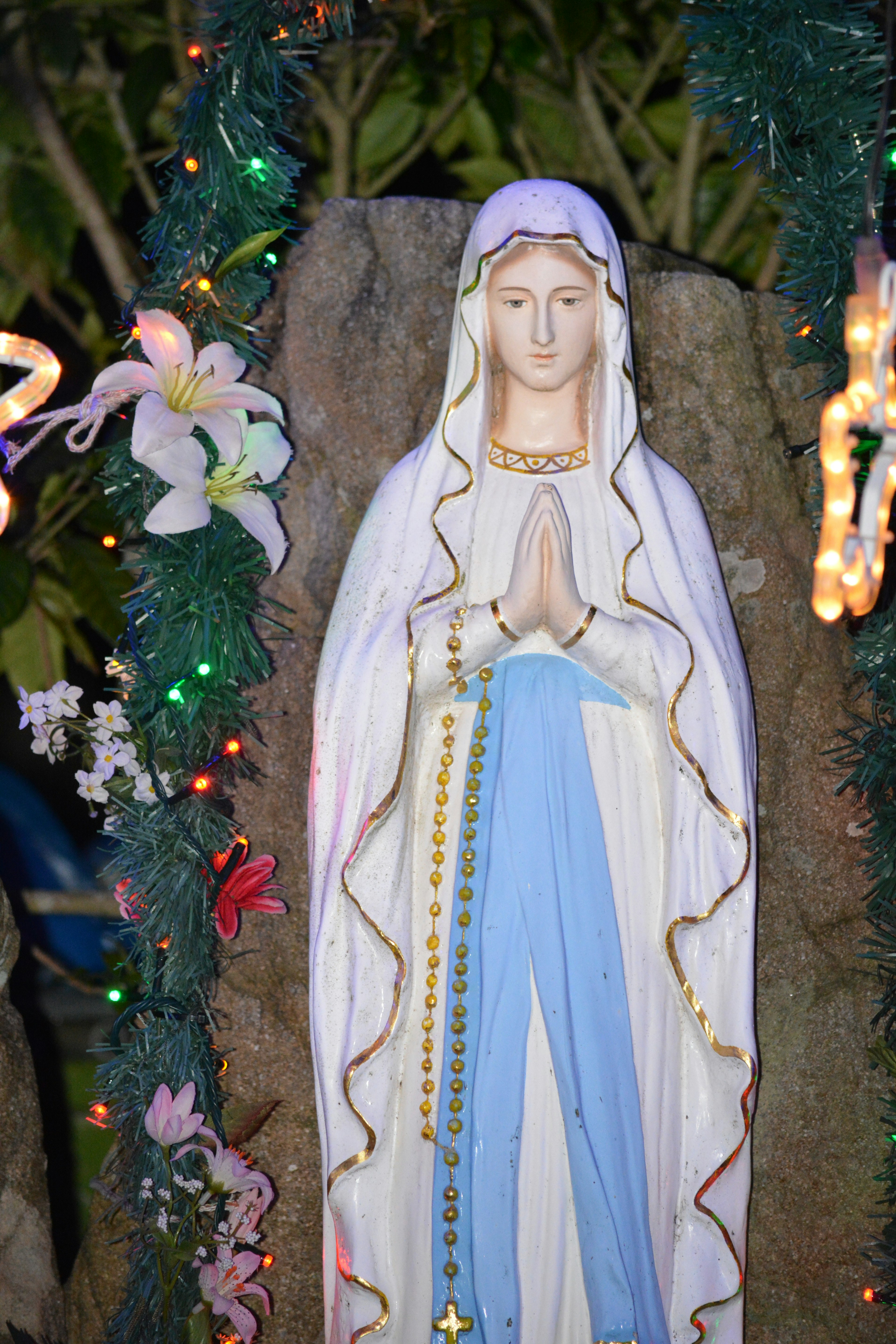 Statue of Mary in blue robe with hands clasped surrounded by flowers and lights