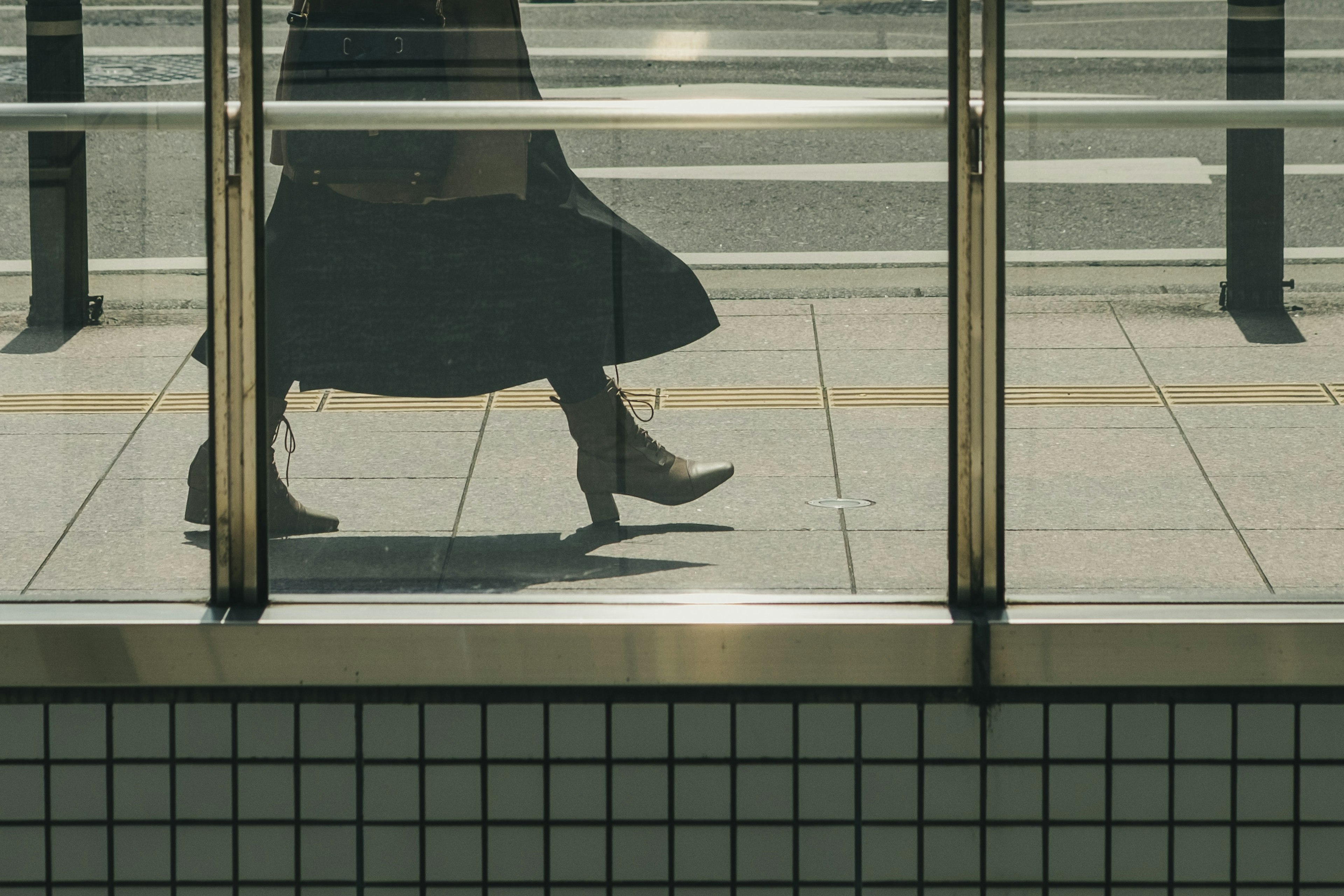 A shadow of a person walking in front of transparent glass wearing a black skirt and boots