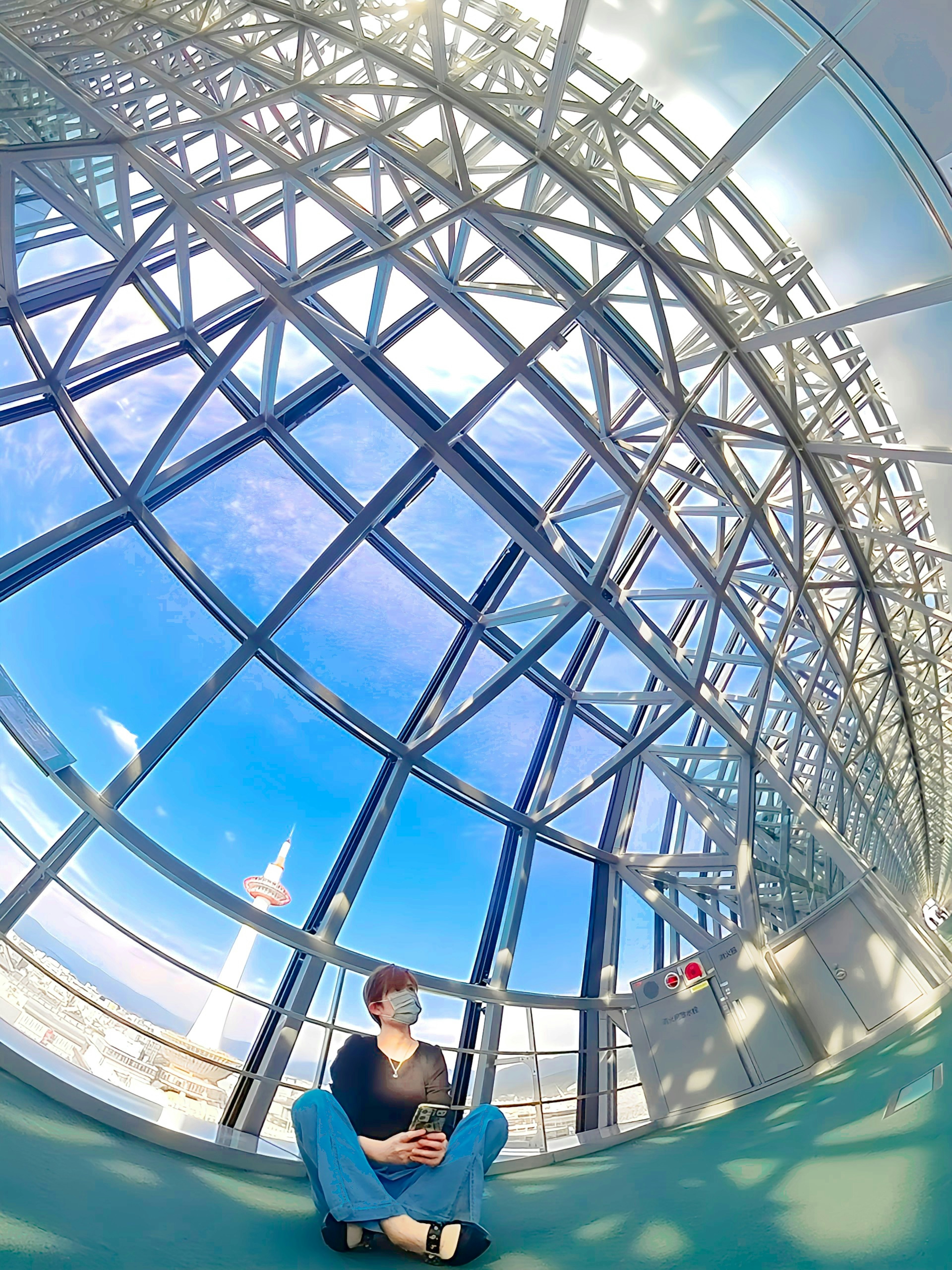 Person sitting inside a large glass structure wearing blue pants and a black top visible blue sky outside