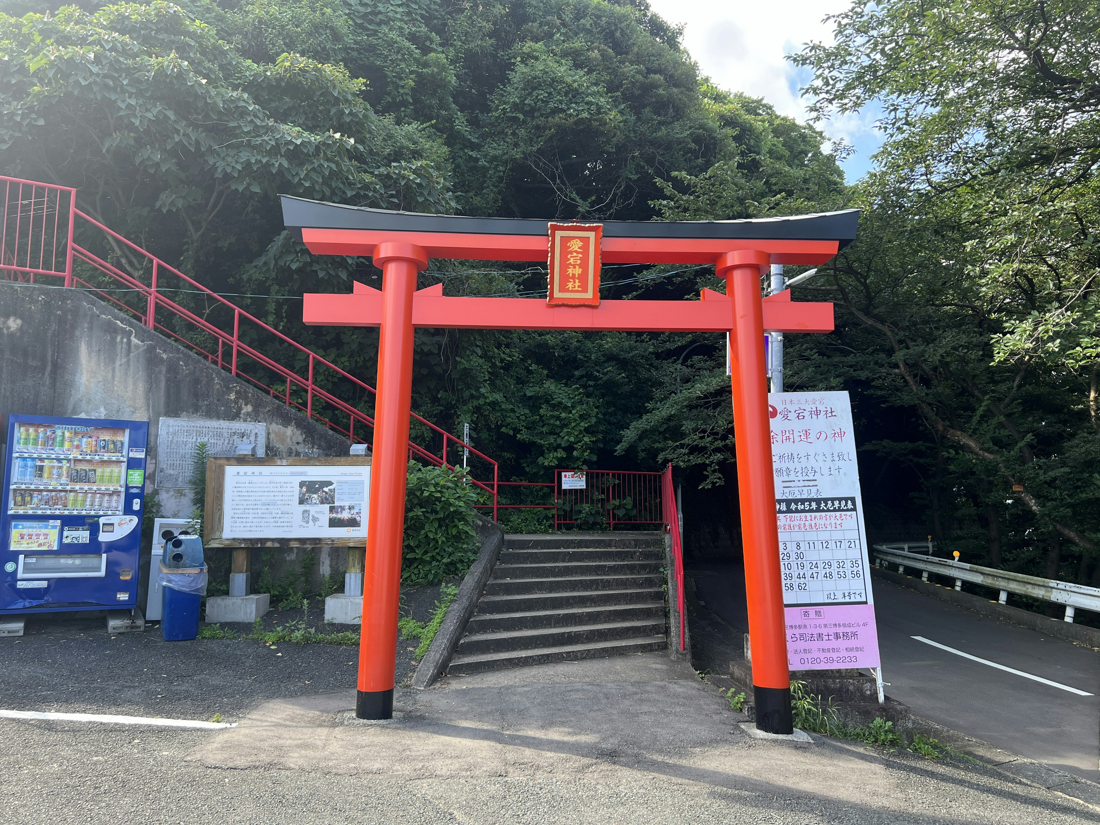 Eingang mit einem roten Torii-Tor, das zu Treppen führt, umgeben von grünen Bäumen