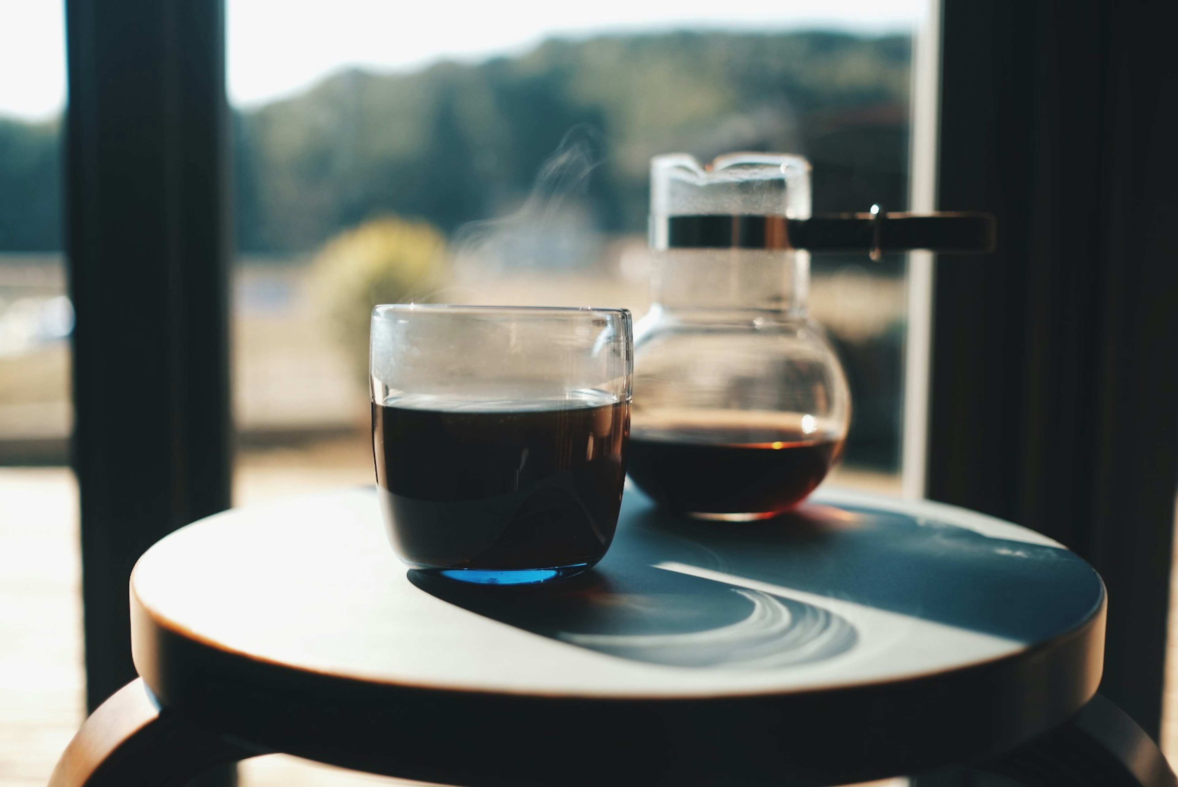 A glass of coffee and a coffee pot placed by the window