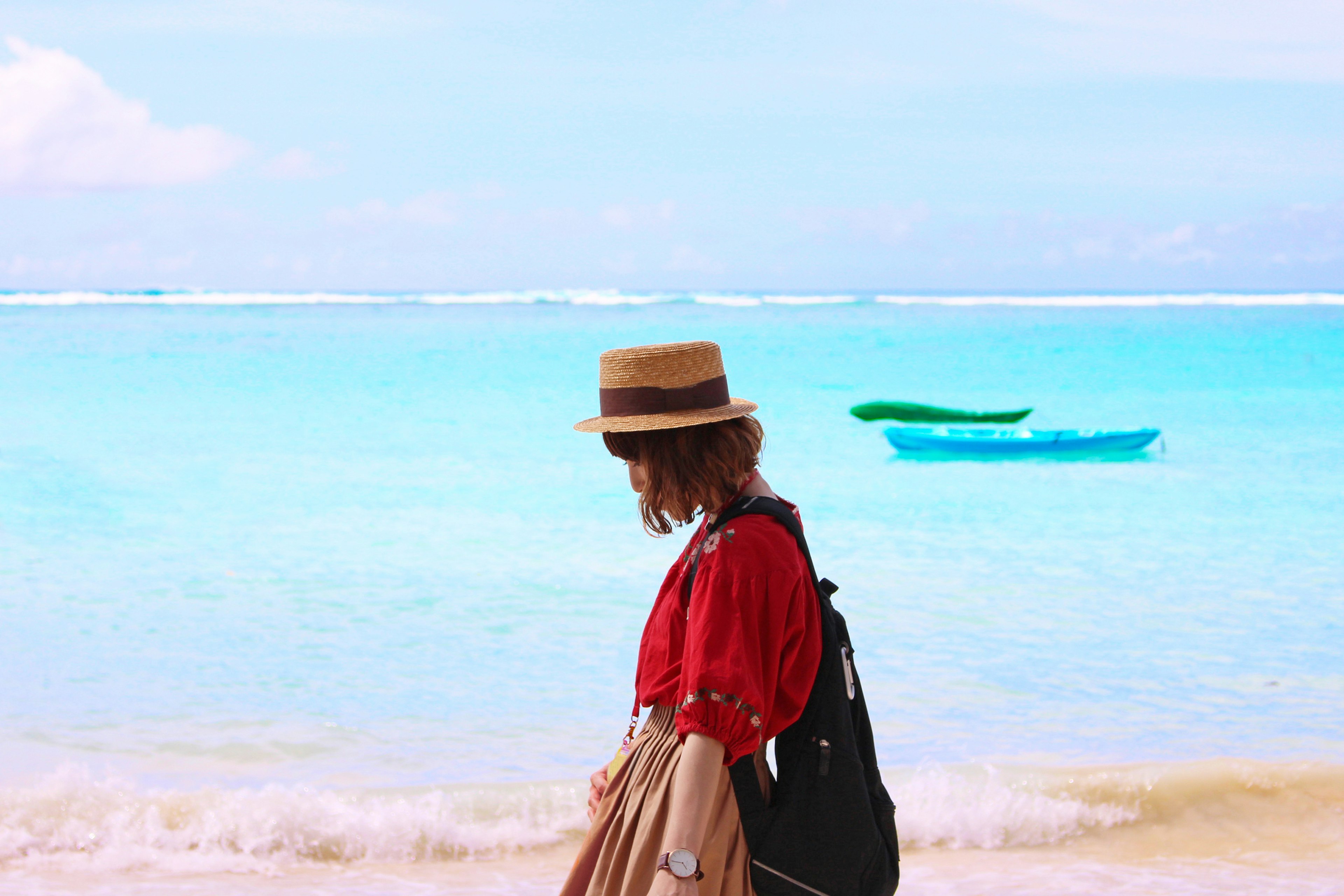 ผู้หญิงใส่หมวกเดินอยู่ริมชายหาดมีน้ำสีเขียวมรกตและเรือคายัคอยู่ในพื้นหลัง