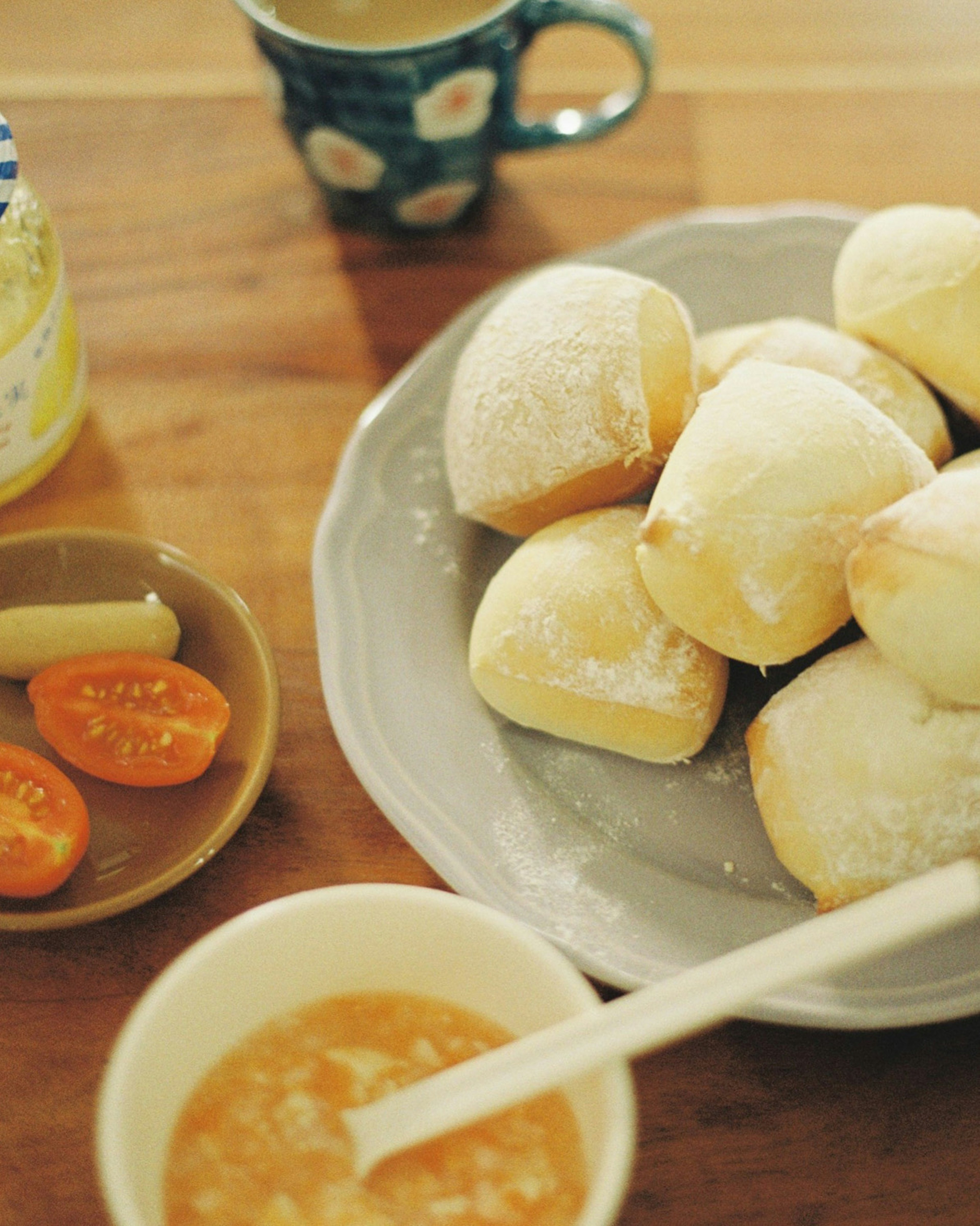 A plate of fluffy bread rolls with a bowl of sauce and a small dish of sliced tomatoes next to a coffee cup