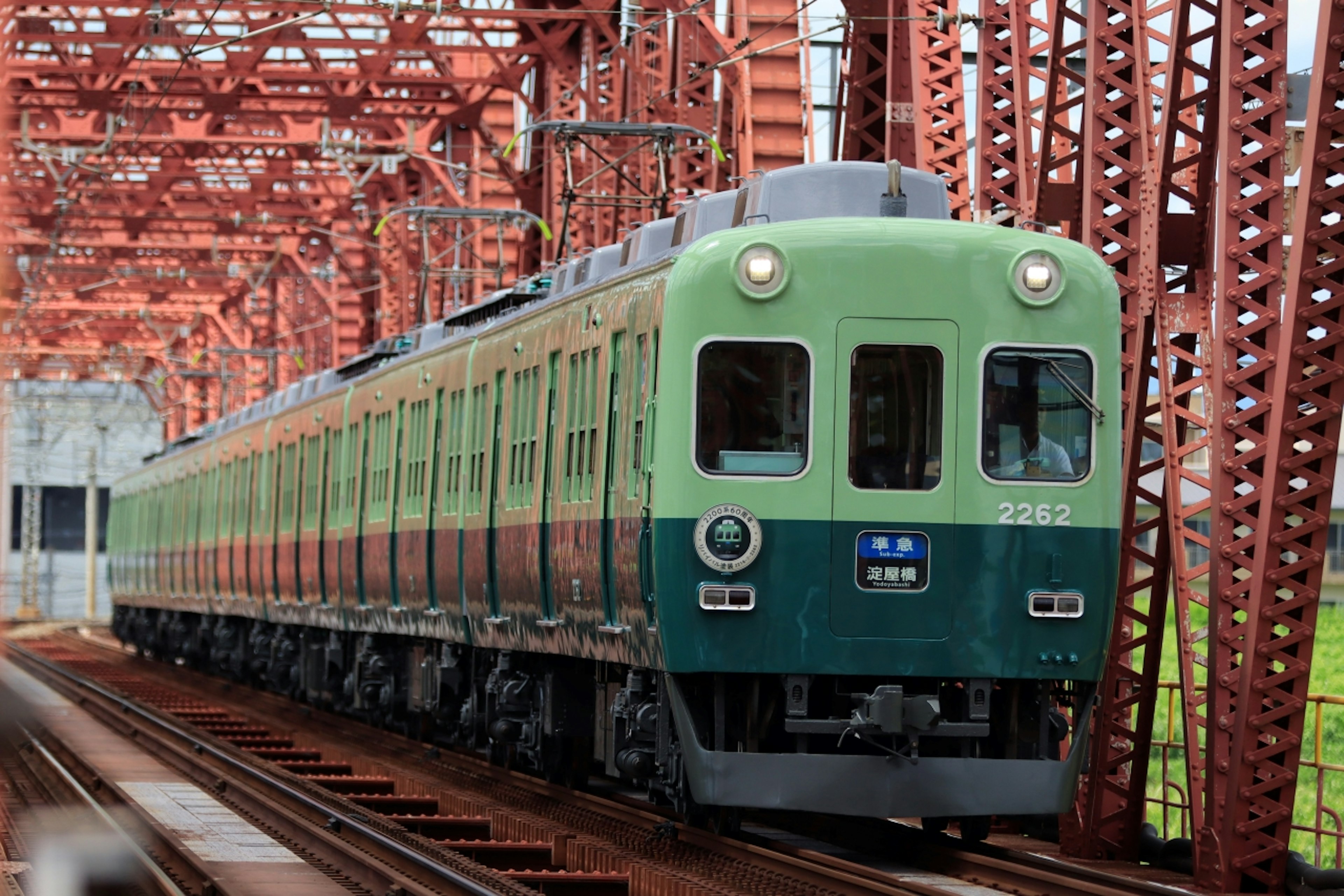 A green and blue commuter train crossing a red iron bridge