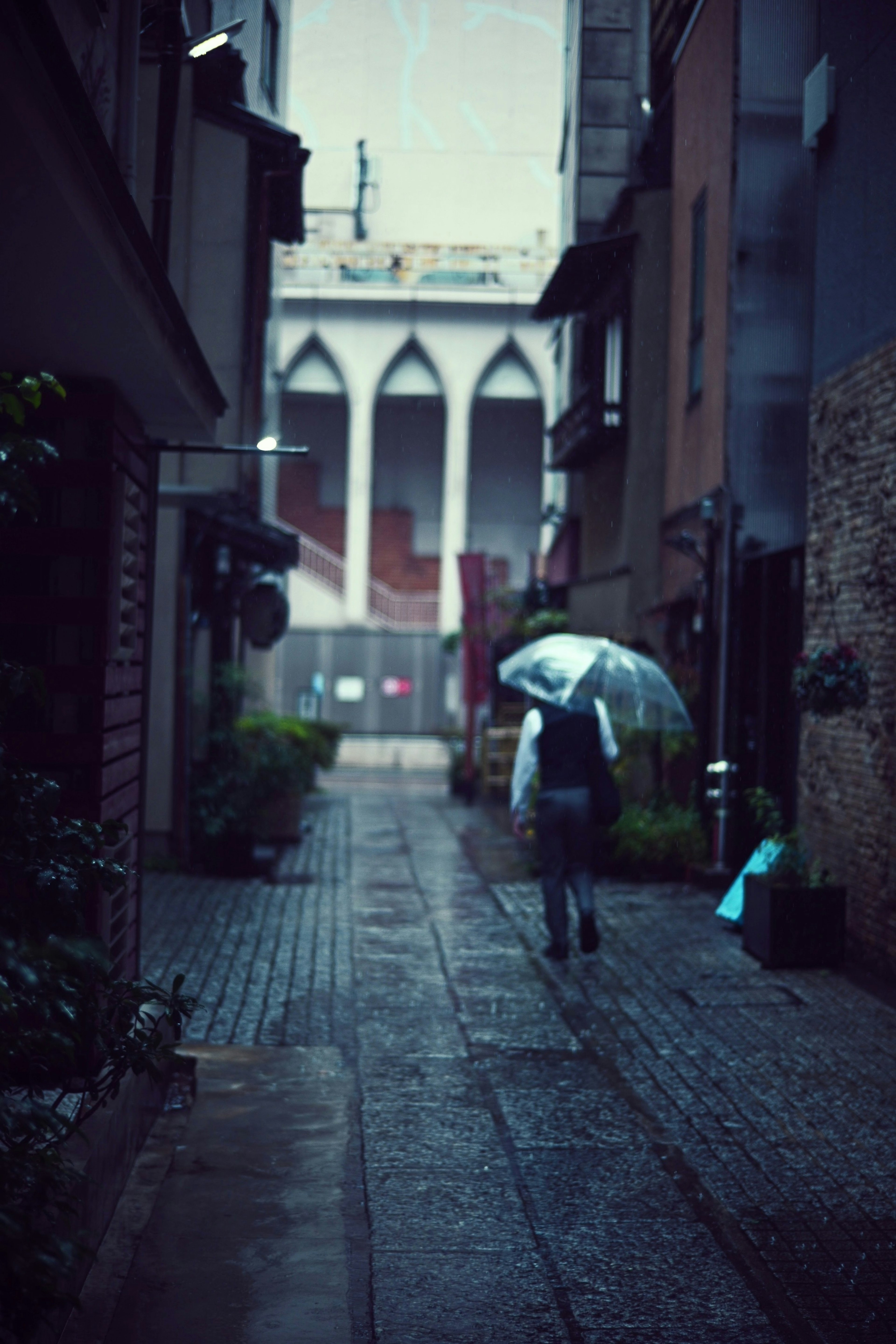 雨の中、傘を持った人が狭い路地を歩いている背景に印象的な建物が見える