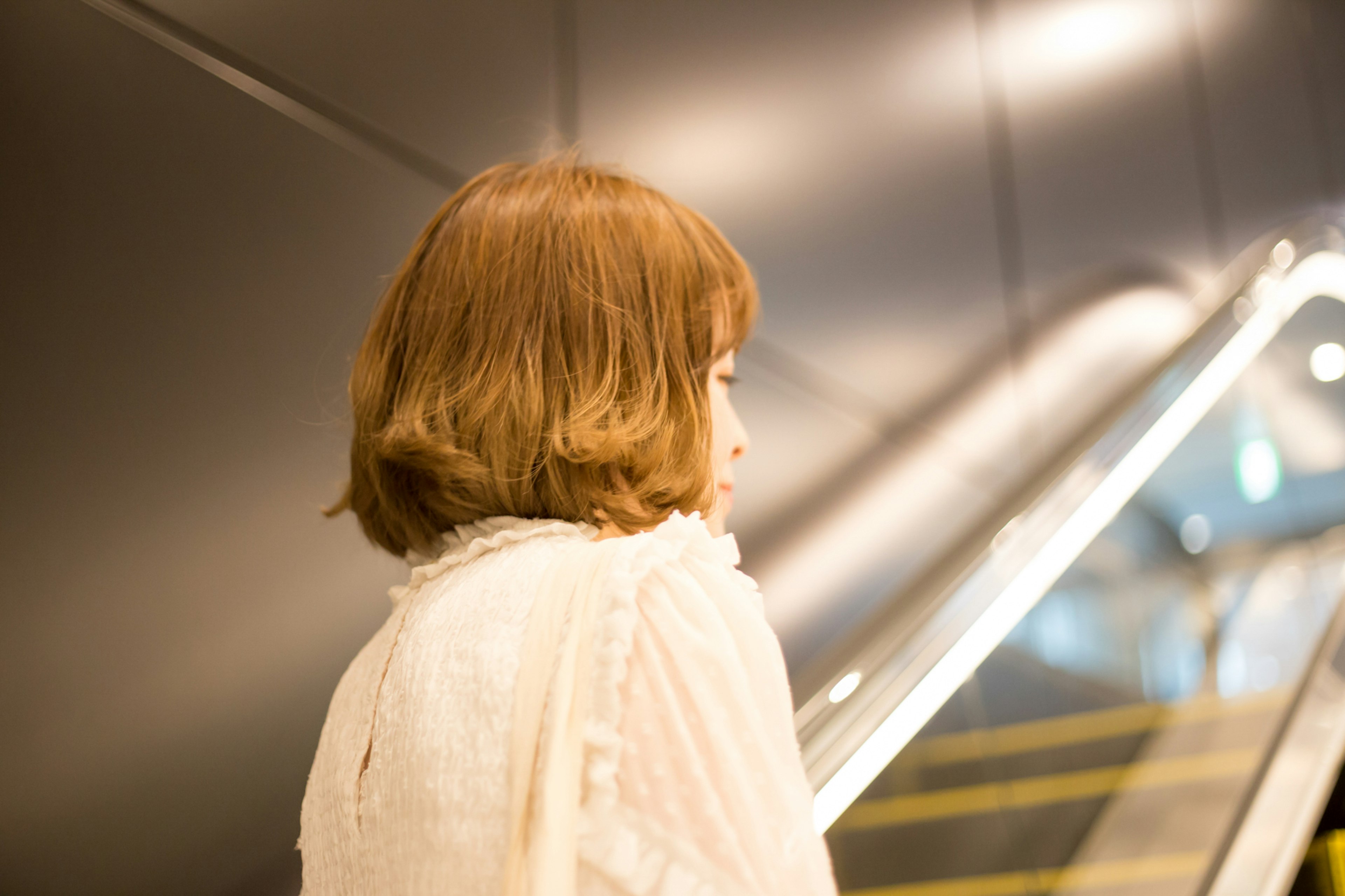 A woman standing near an escalator seen from behind with brown bobbed hair