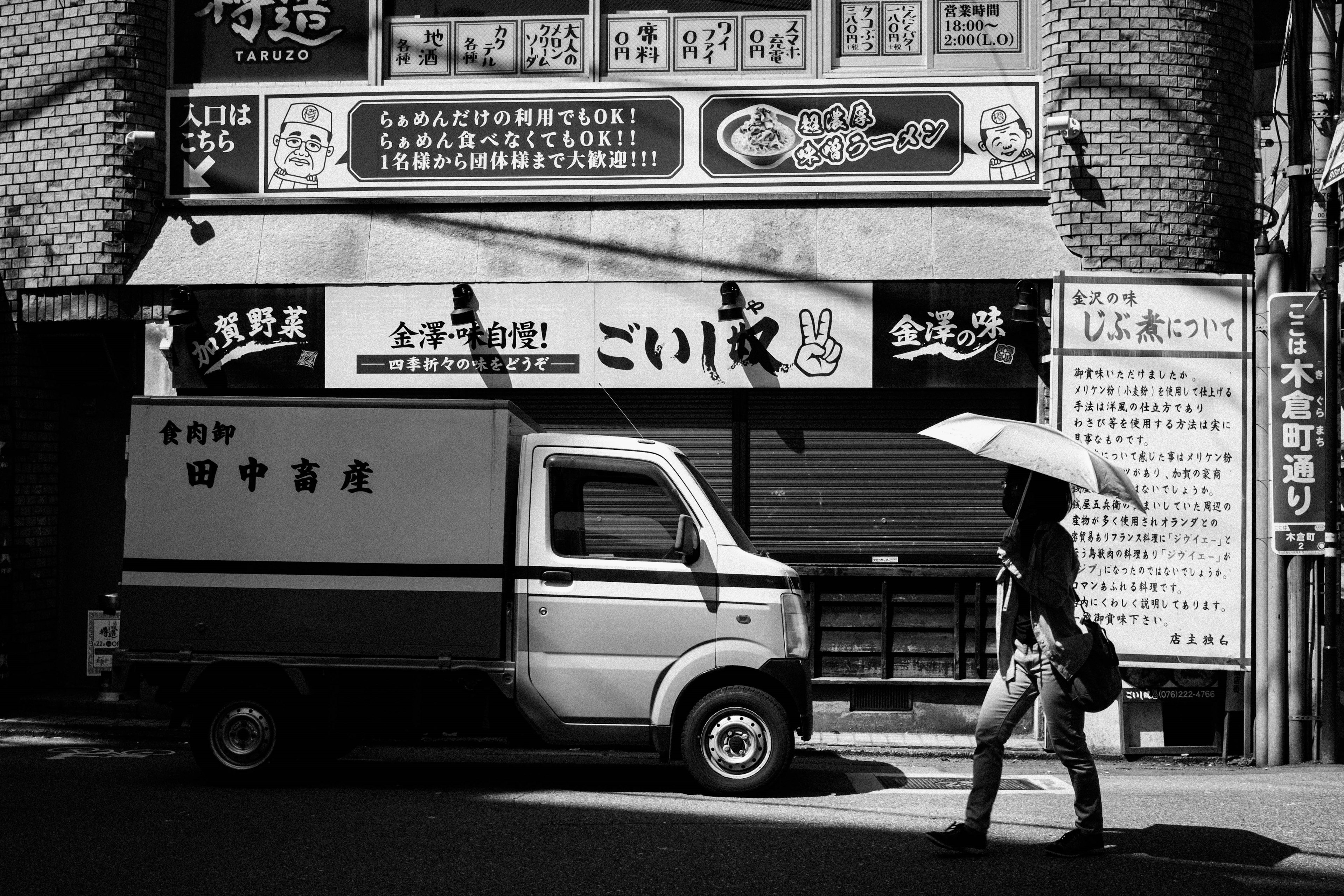 Una escena de calle en blanco y negro con una persona caminando con un paraguas y una camioneta de entrega en un área comercial