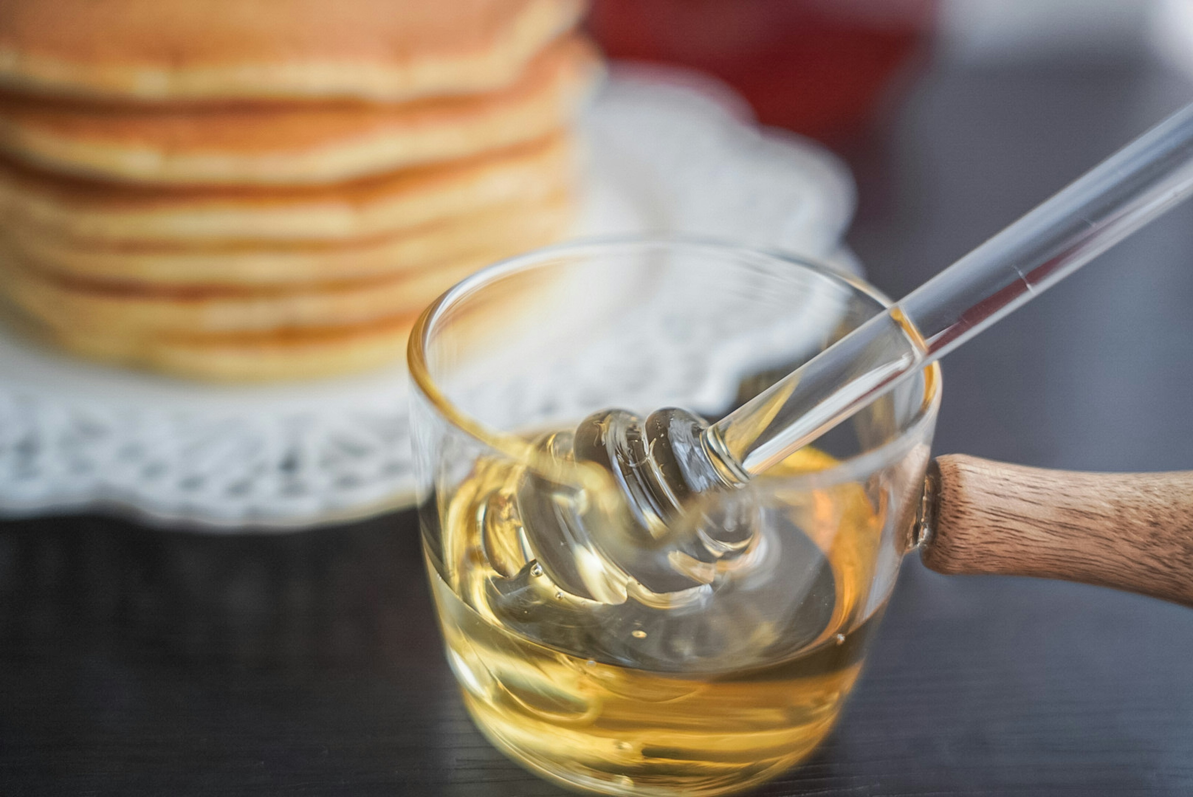 Madu dalam piring kaca dengan sendok madu dan pancake di latar belakang