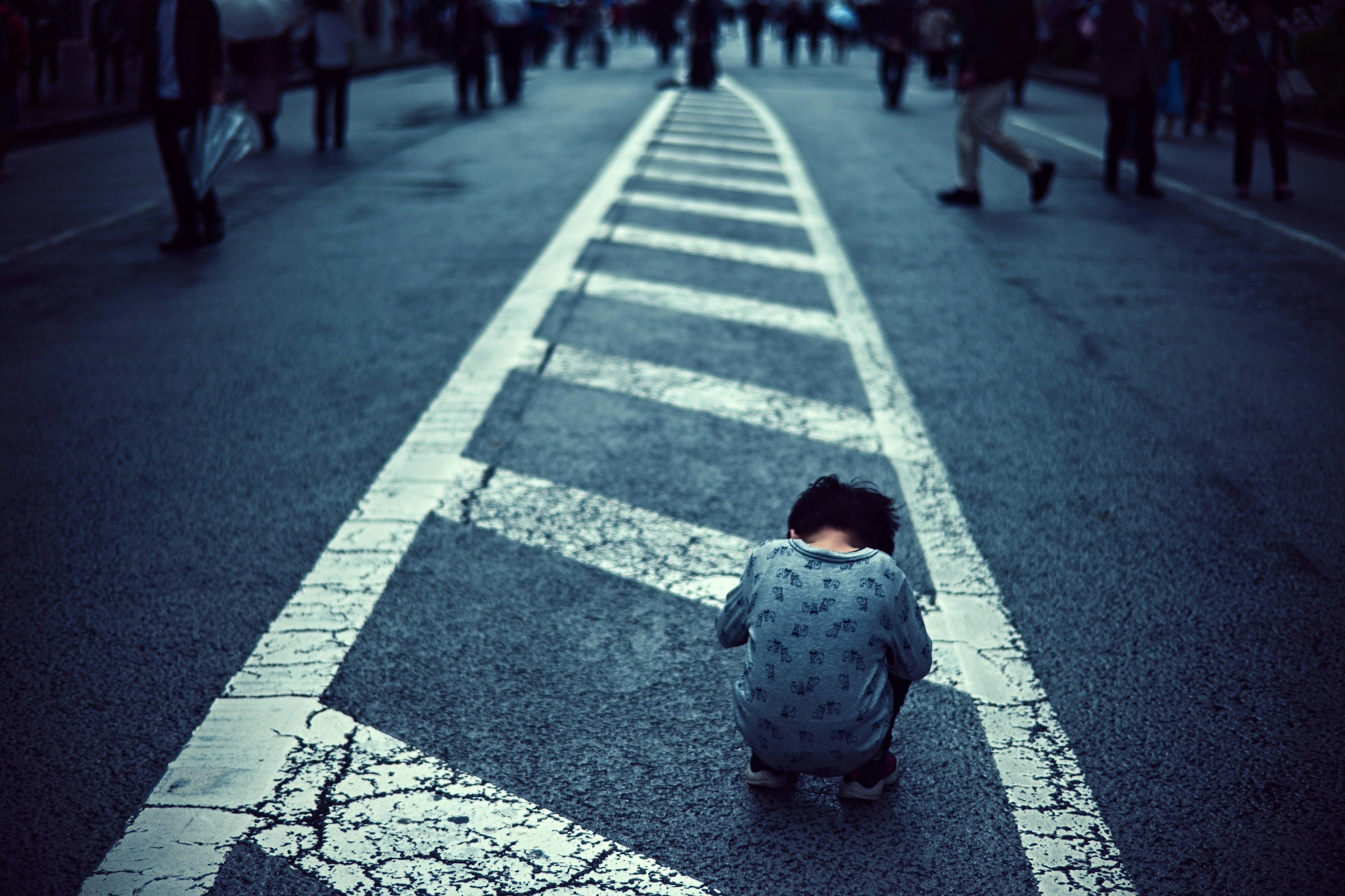 A child squatting on a cracked white crosswalk with pedestrians in the background