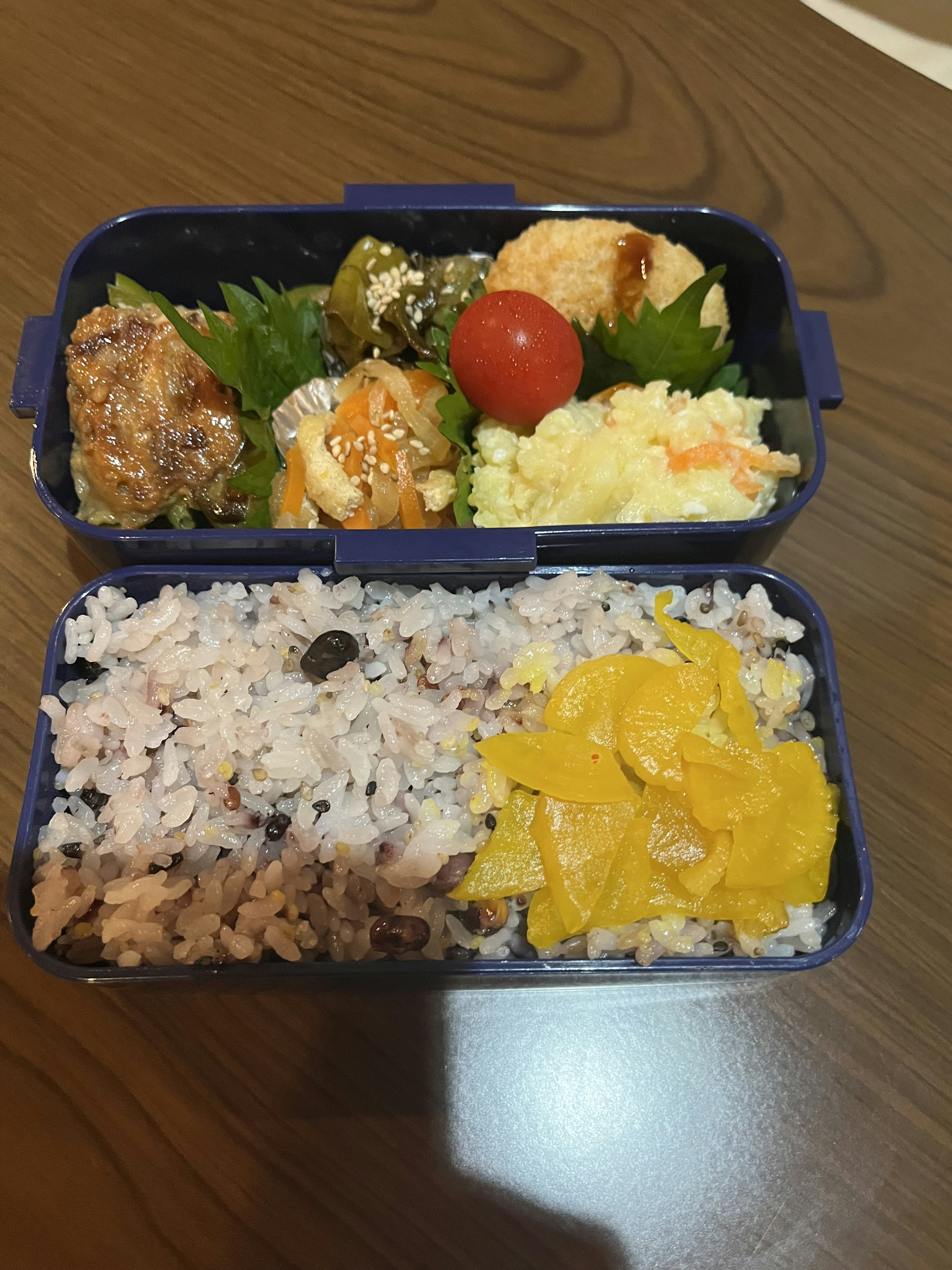 A bento box featuring chicken, vegetables, egg salad, and cherry tomatoes on the top section with multigrain rice and mango slices on the bottom