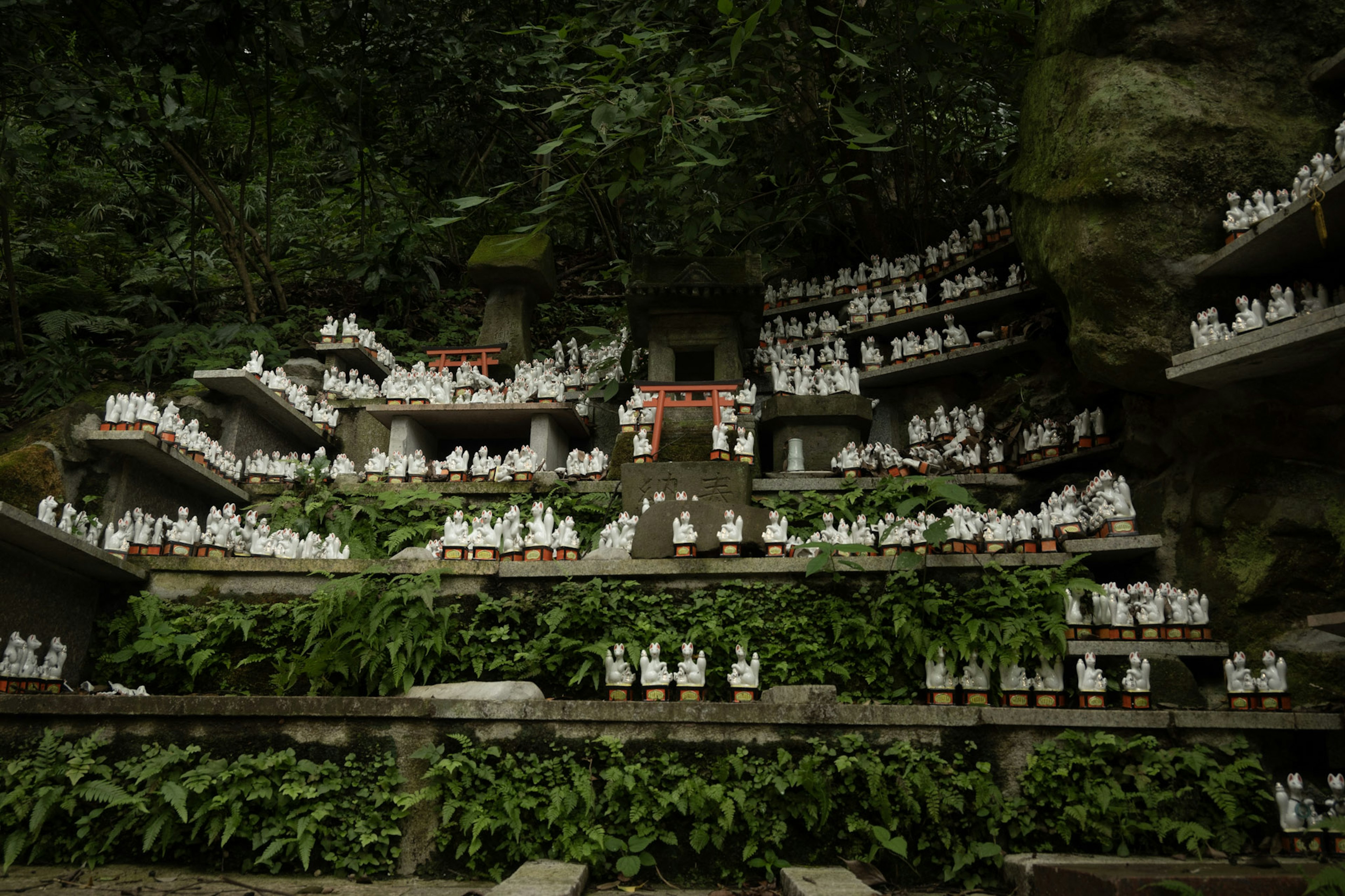 Small white statues arranged on stone shelves surrounded by greenery with a small red shrine