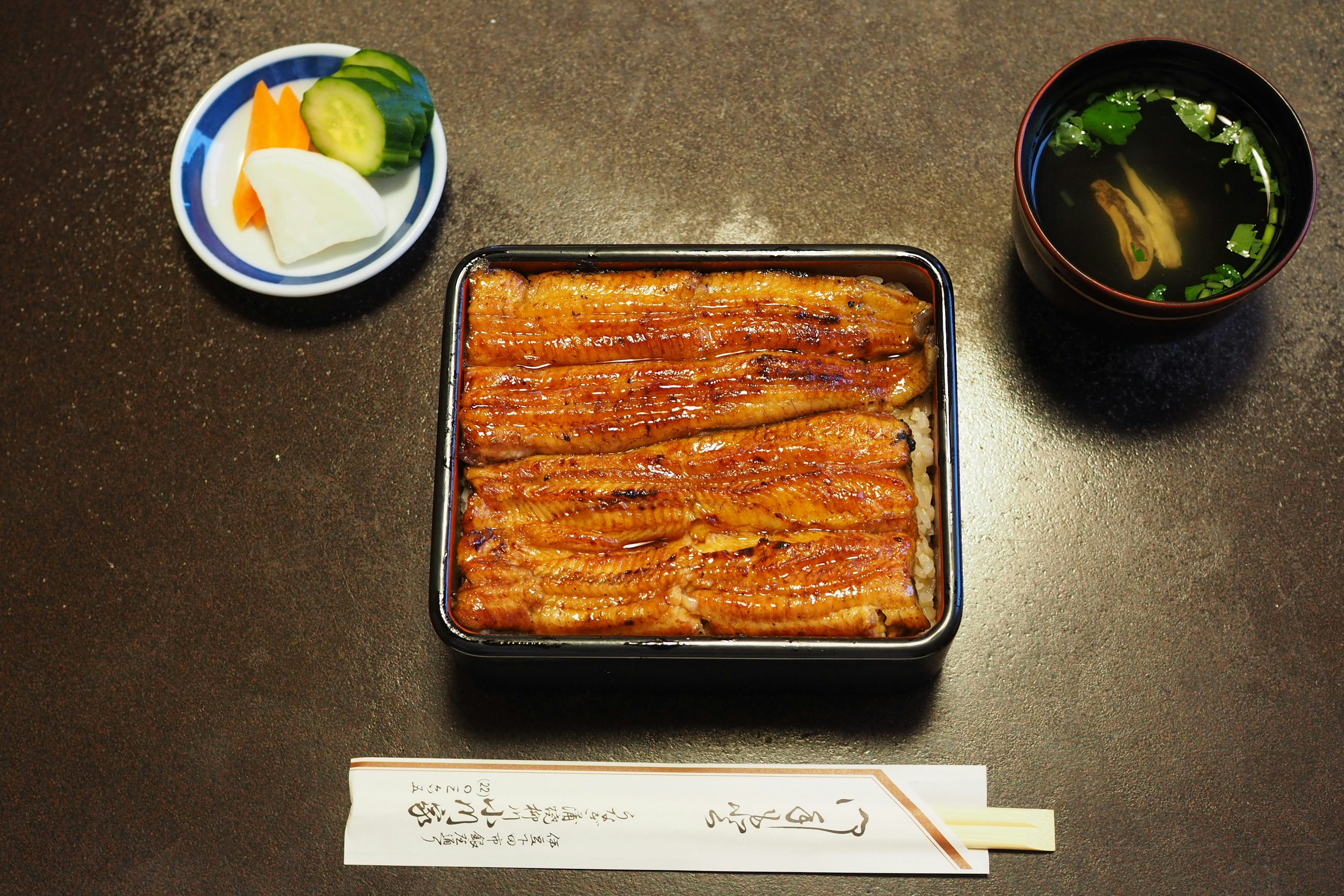 A black box containing grilled eel rice with pickled vegetables and miso soup on the side