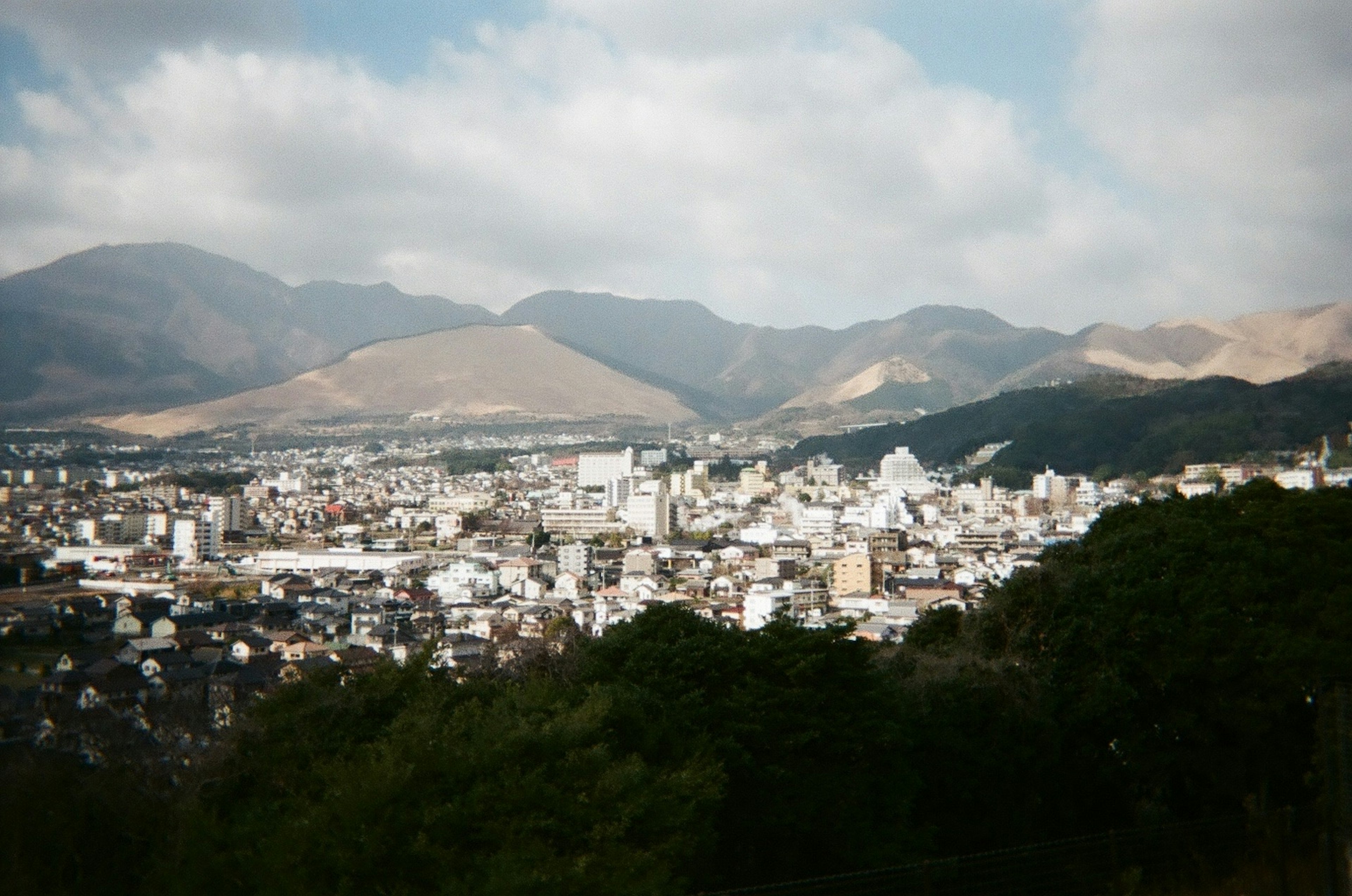 山々に囲まれた都市の風景で、白い建物と緑の木々が見える