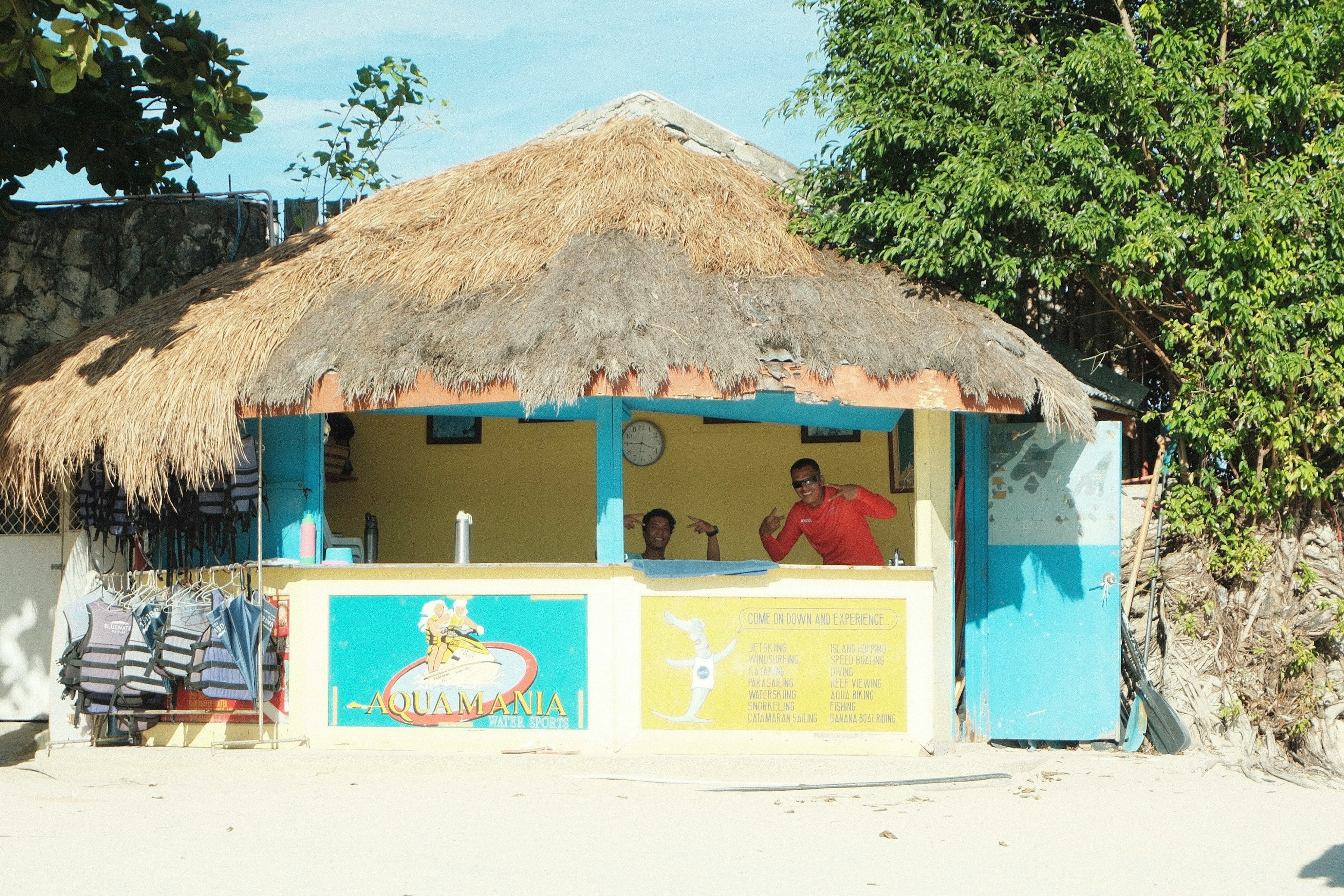 Außenansicht einer Strandhütte mit Strohdach und blauen Wänden mit zwei Mitarbeitern am Counter