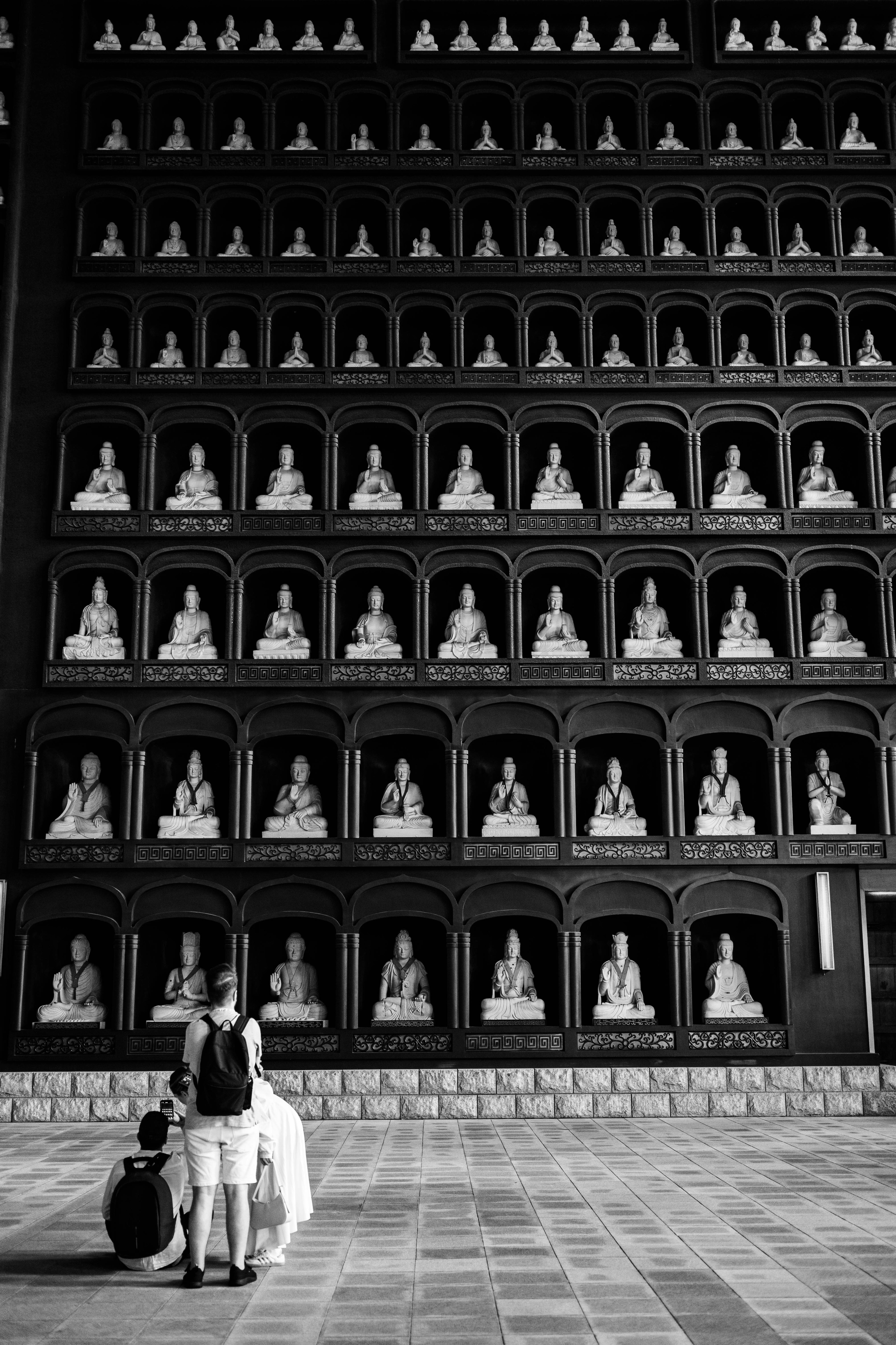 Two individuals praying in front of a wall filled with Buddha statues