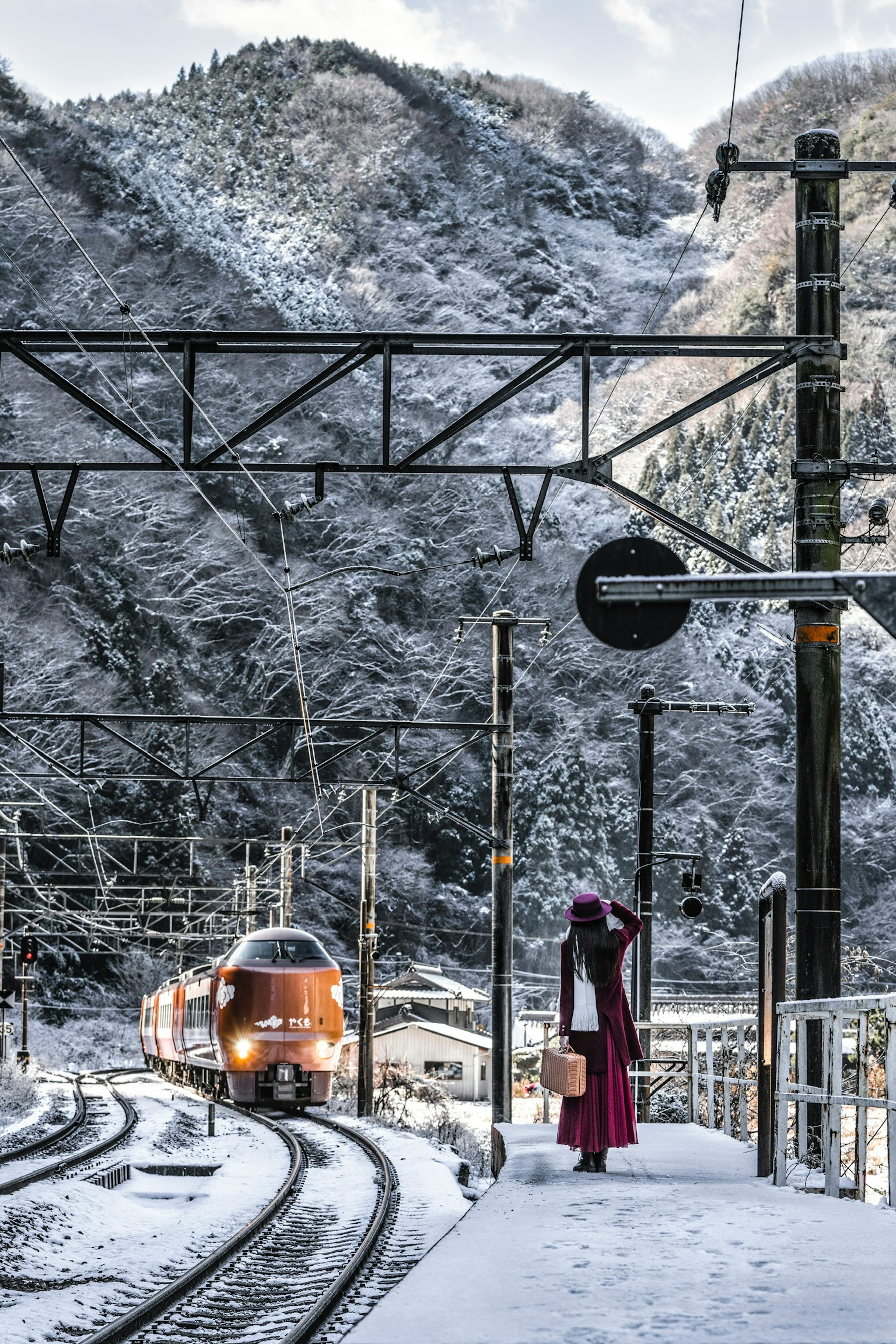 Train arriving at a snowy station with mountains in the background and a person in red clothing