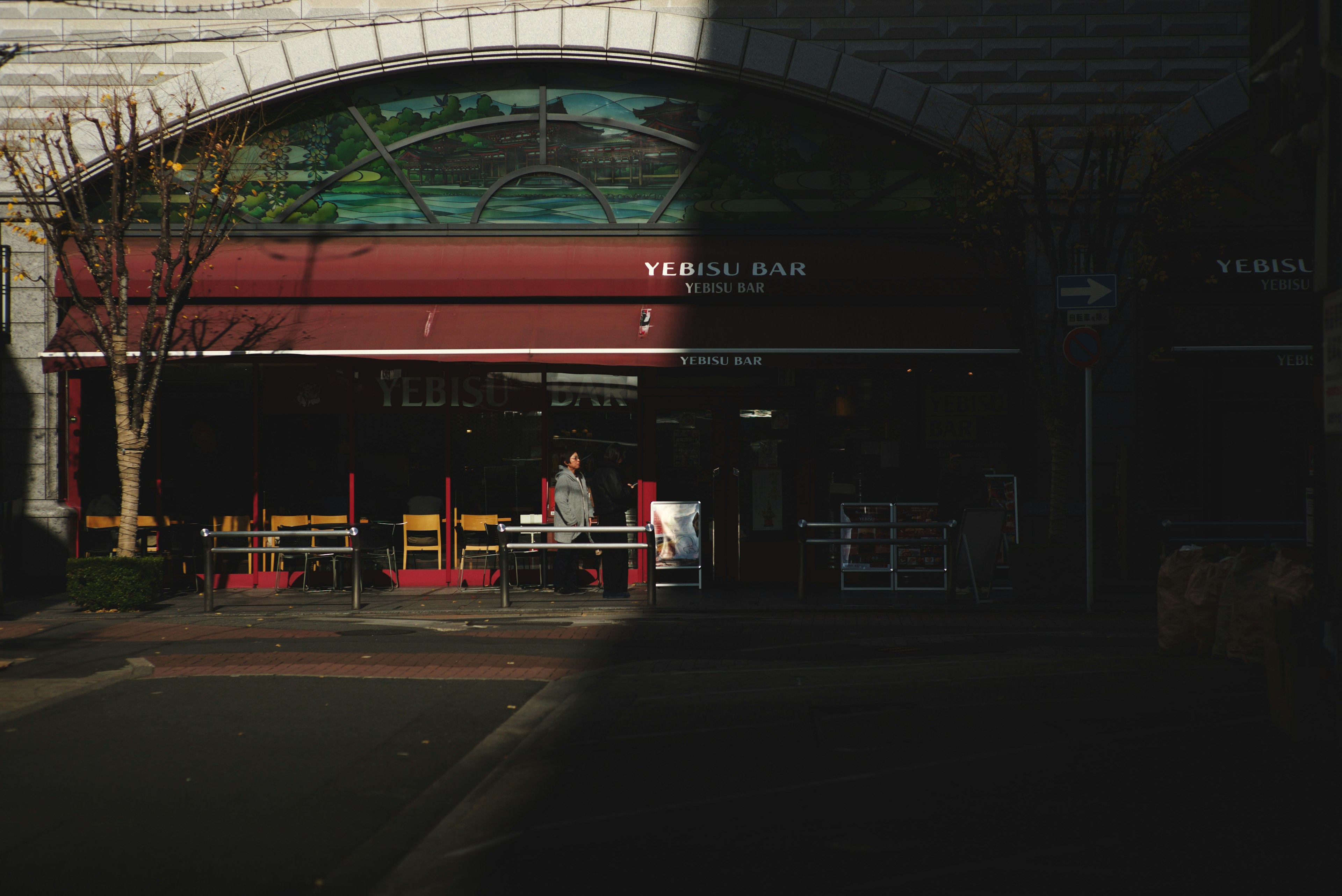 Extérieur d'un café avec des chaises rouges dans l'ombre et un toit en verre lumineux