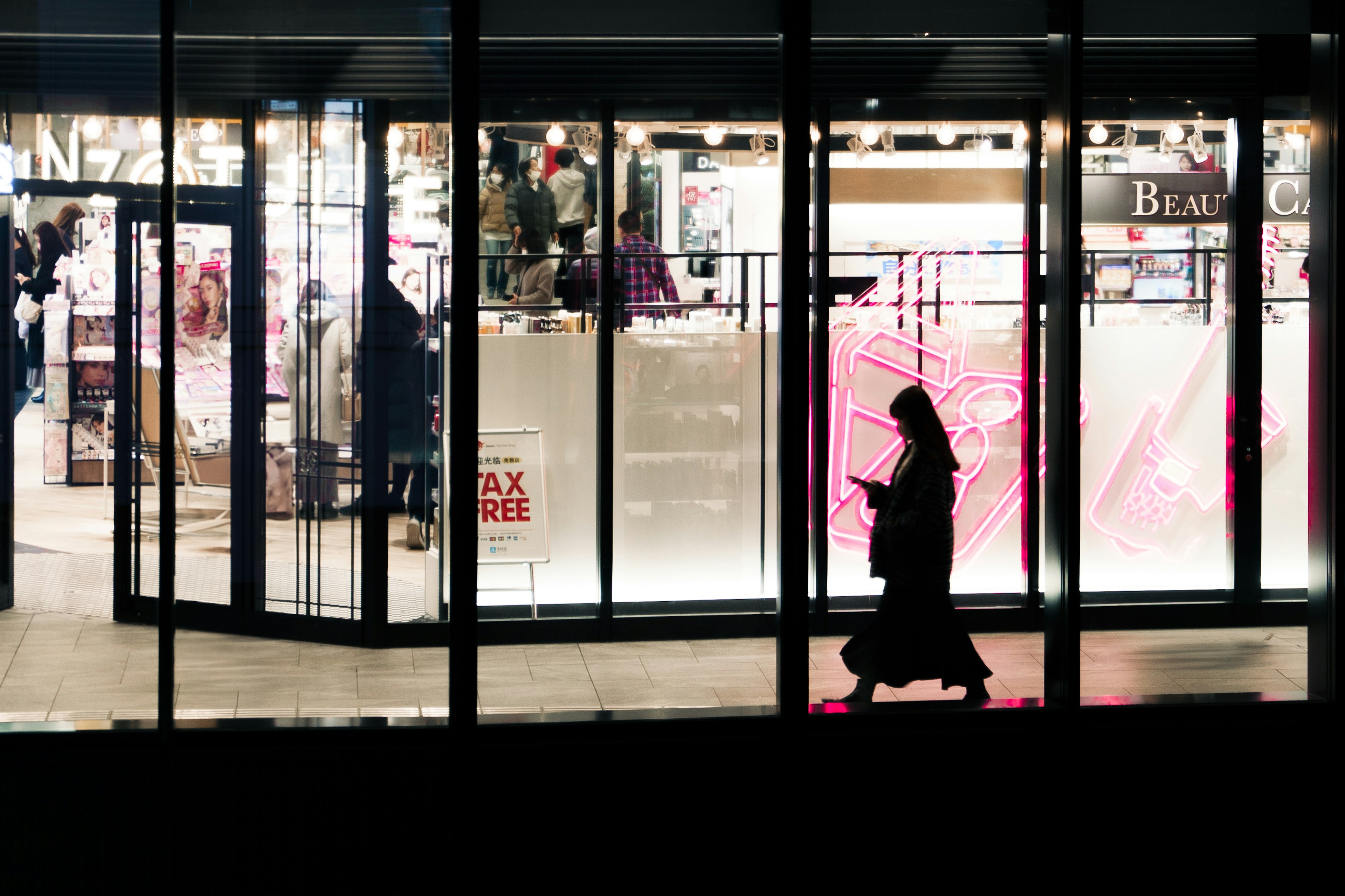 Silhouette einer Frau, die mit einem Smartphone in einer schwach beleuchteten Straßenecke mit hellen Ladeninterieurs geht