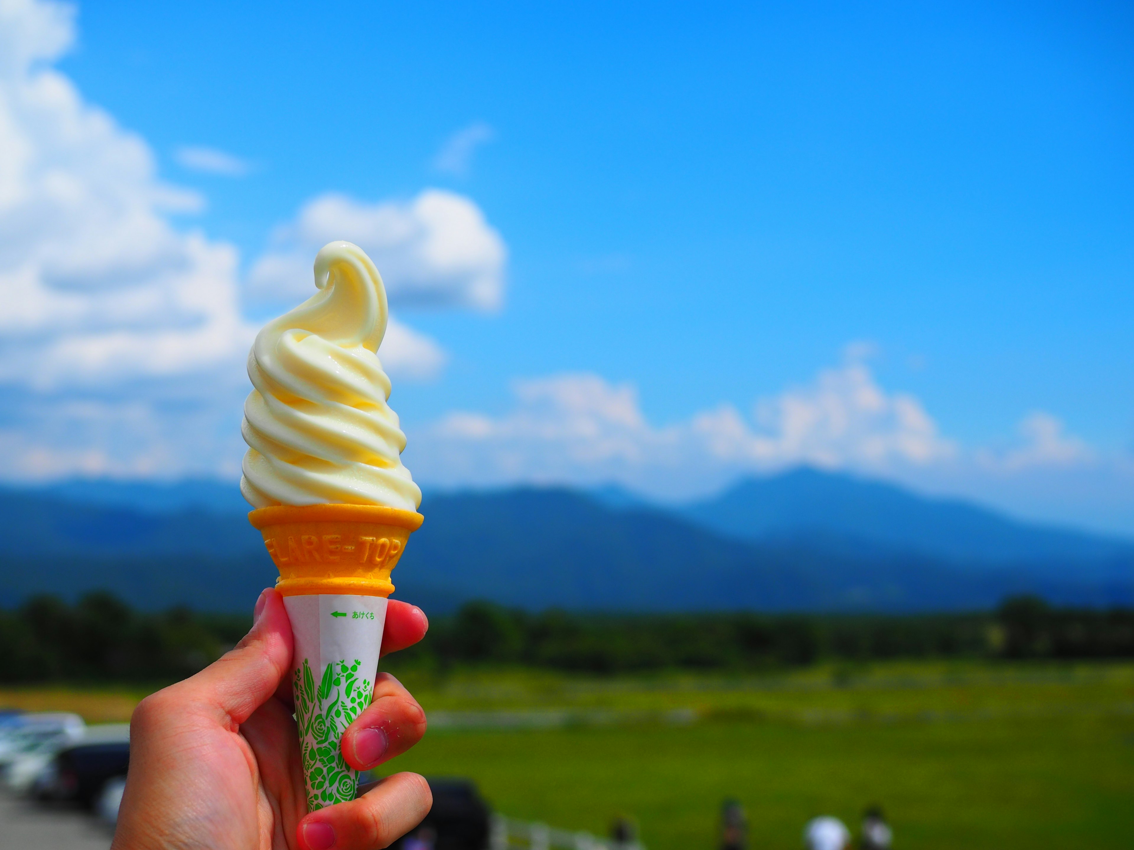 Hand holding soft serve ice cream cone against blue sky and mountains