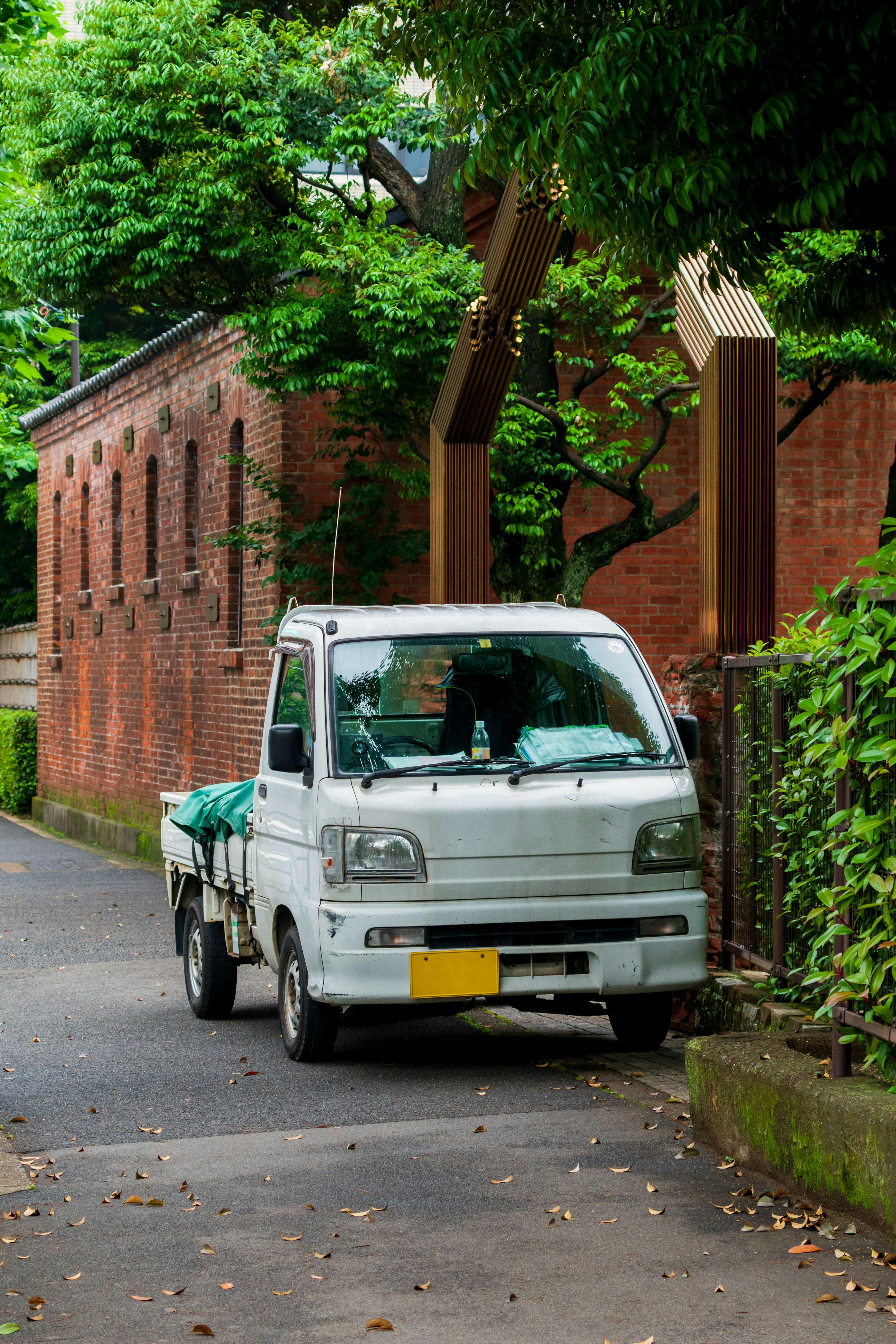 白い軽トラックが緑の木々と赤レンガの壁の間に停まっている