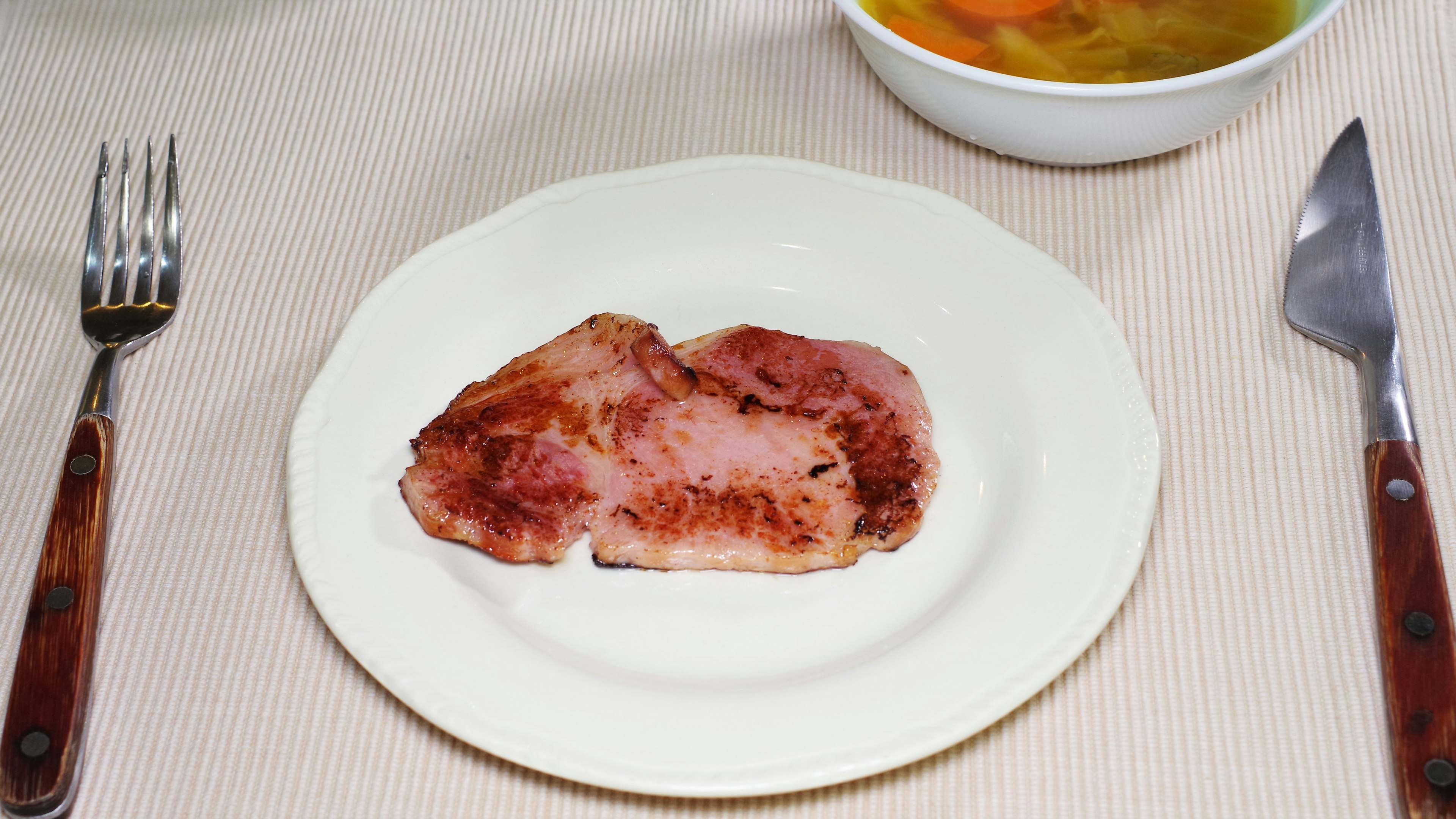 A slice of cooked meat on a plate with a bowl of soup in the background