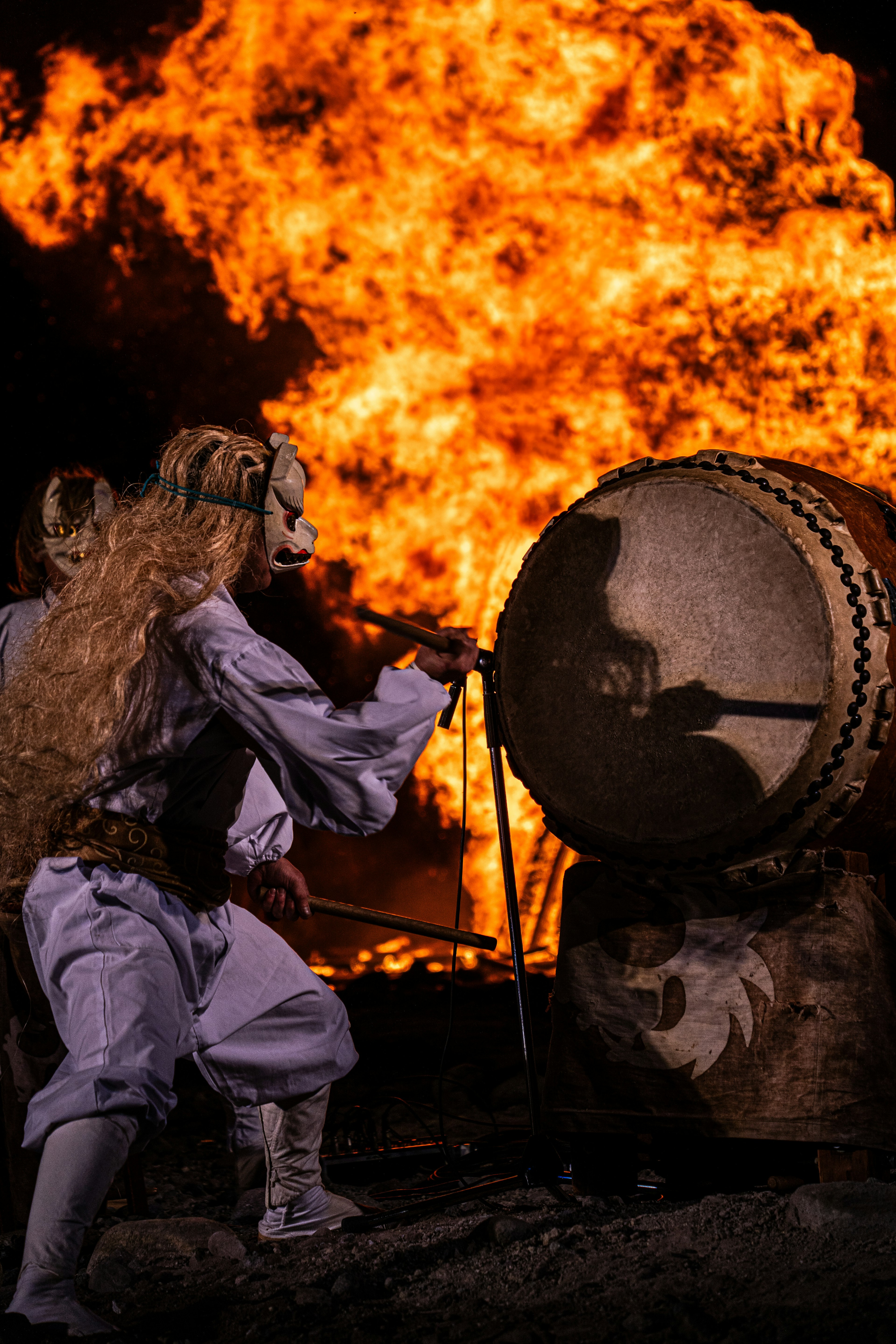 Seorang penampil dengan pakaian tradisional memainkan drum di depan api besar Sosok berambut panjang dengan latar belakang dramatis