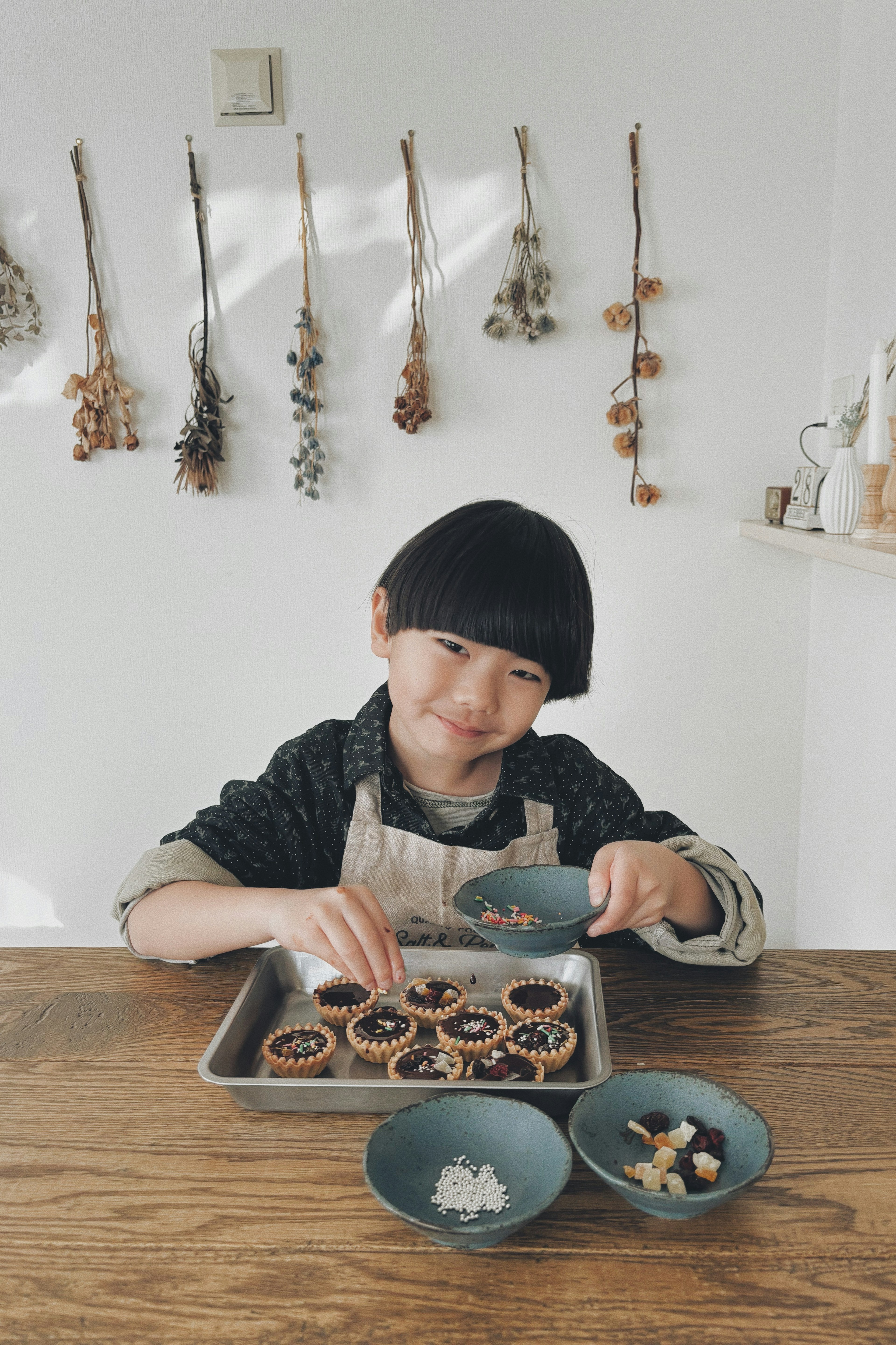 Bambino che decora biscotti su un tavolo Ciotole carine con ingredienti davanti Fiori secchi appesi sullo sfondo